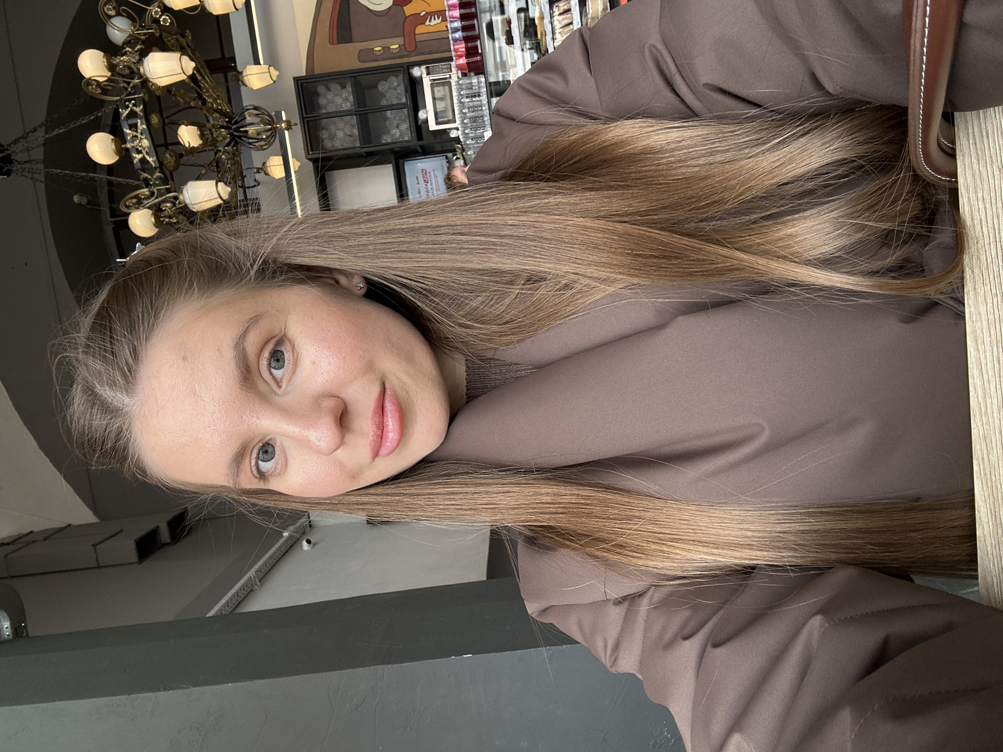 A woman with fair skin and long, straight, light-brown hair is indoors, wearing a brown puffy jacket. She is seated at a table, the edge of which is visible to the right. In the background, a chandelier hangs from the ceiling, and a display of various objects is visible. The location is Moscow, Russia.