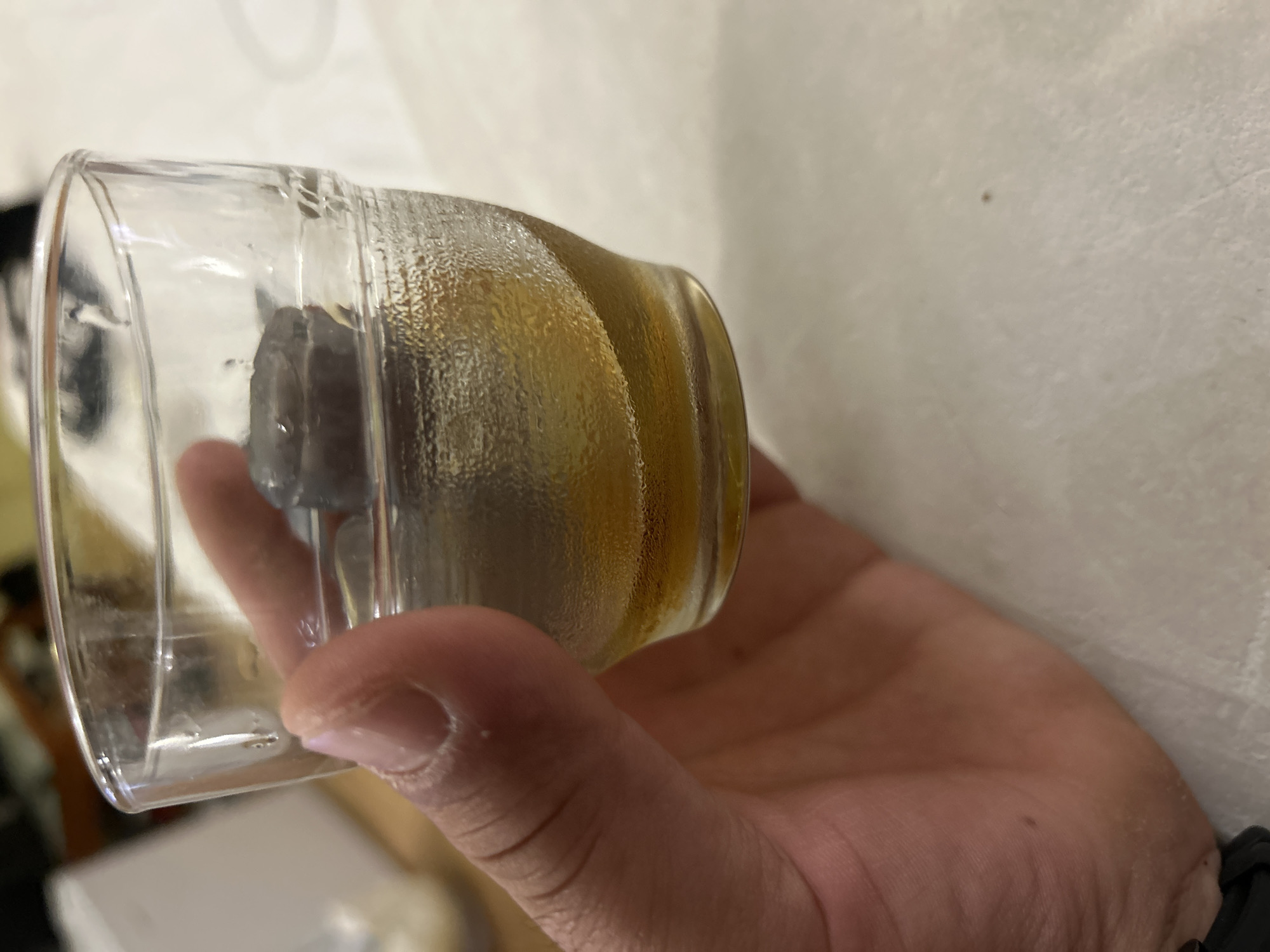 A clear glass with ice and amber-colored liquid is being held by a person's hand. Condensation is visible on the outside of the glass. The background is out of focus, showing a white wall. The location, Sveti Vlas, Bulgaria, is not discernible from the visual content of the image.