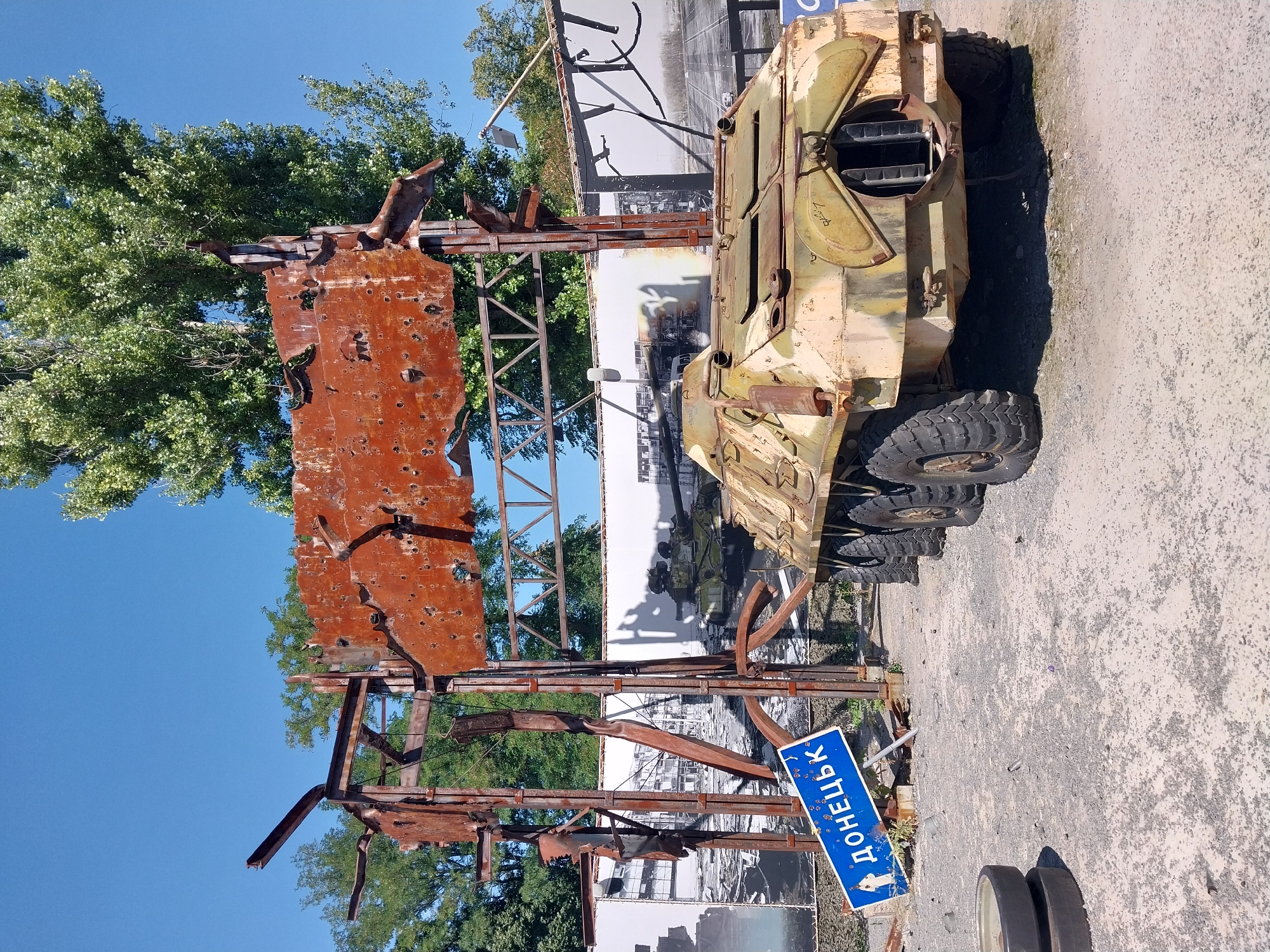 The image captures an outdoor scene in Dnipro, Ukraine, featuring a damaged Soviet-era armored personnel carrier (APC) and a rusted metal structure resembling a gateway or archway, both bearing evidence of significant impact, likely from shelling or explosions. The APC, painted in a camouflage pattern of beige and green, is positioned to the right of the frame, facing towards the right. It exhibits signs of wear and tear, with some panels appearing warped or detached. The metal structure, prominently displayed in the center, is heavily corroded and perforated with numerous holes, indicative of bullet or shrapnel damage.
In front of the metal structure, a blue road sign with white lettering reads "ДОНЕЦЬК" (Donetsk) in Cyrillic script, and features a white silhouette of a lone figure. The ground is covered with gravel and dirt. Behind the APC and the metal structure, large black and white photographic murals are visible, depicting urban destruction and military vehicles, which appear to be part of a memorial or exhibition. The backdrop consists of lush green trees and a clear blue sky. The overall impression is one of a war-torn landscape preserved as a testament to conflict. The location is specifically identified as the Memorial Complex of the Nadia Museum, situated at the former Dnipro airport.