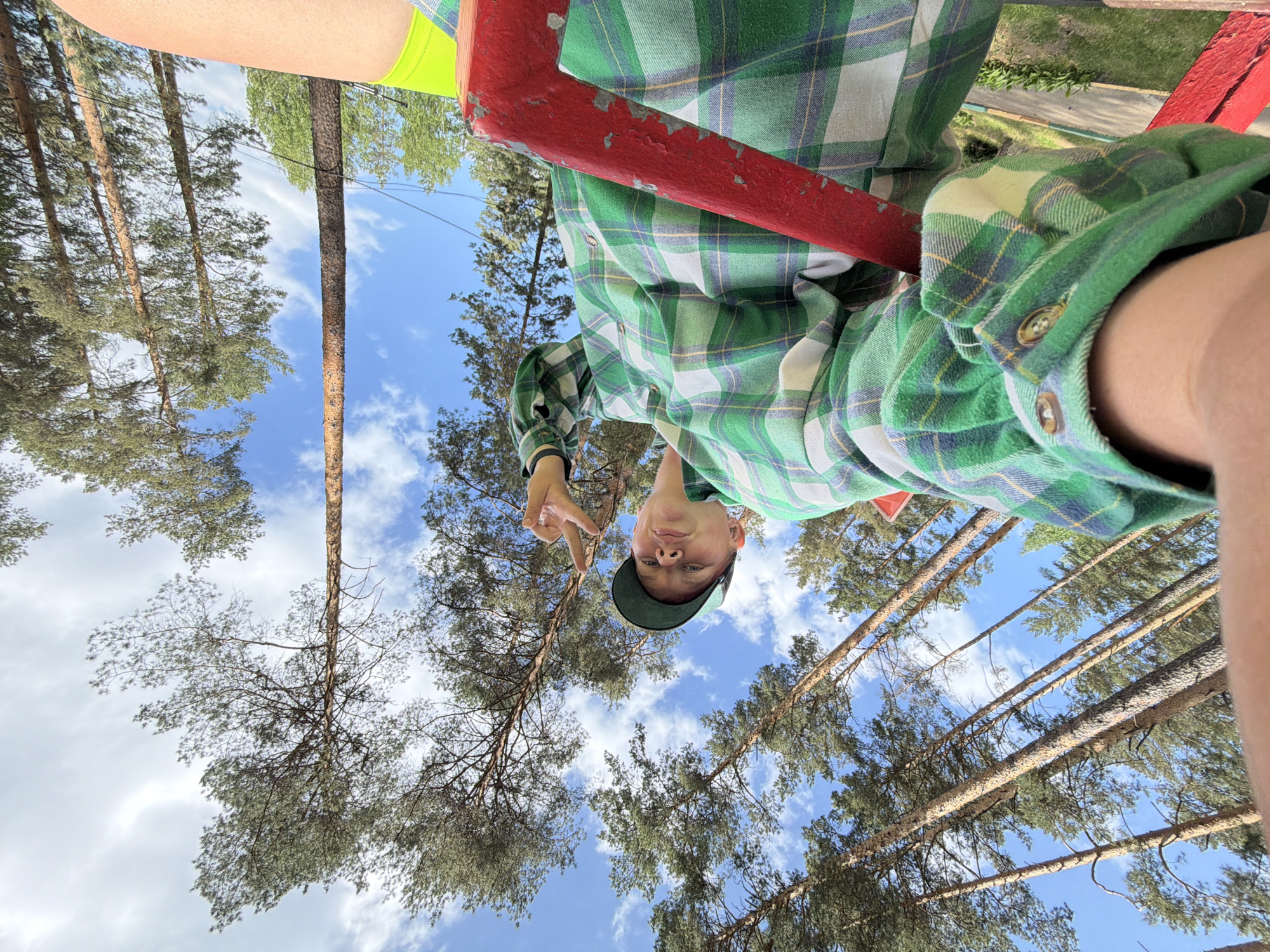 A low-angle view looking up at a person wearing a green plaid shirt and a green cap, who is making a peace sign with their fingers. The person is standing next to a red metal structure. The background shows tall pine trees and a blue sky with scattered clouds. The location appears to be an outdoor area with trees, possibly near Chaykovsky, Russia, given the presence of pine trees and a light blue sky.