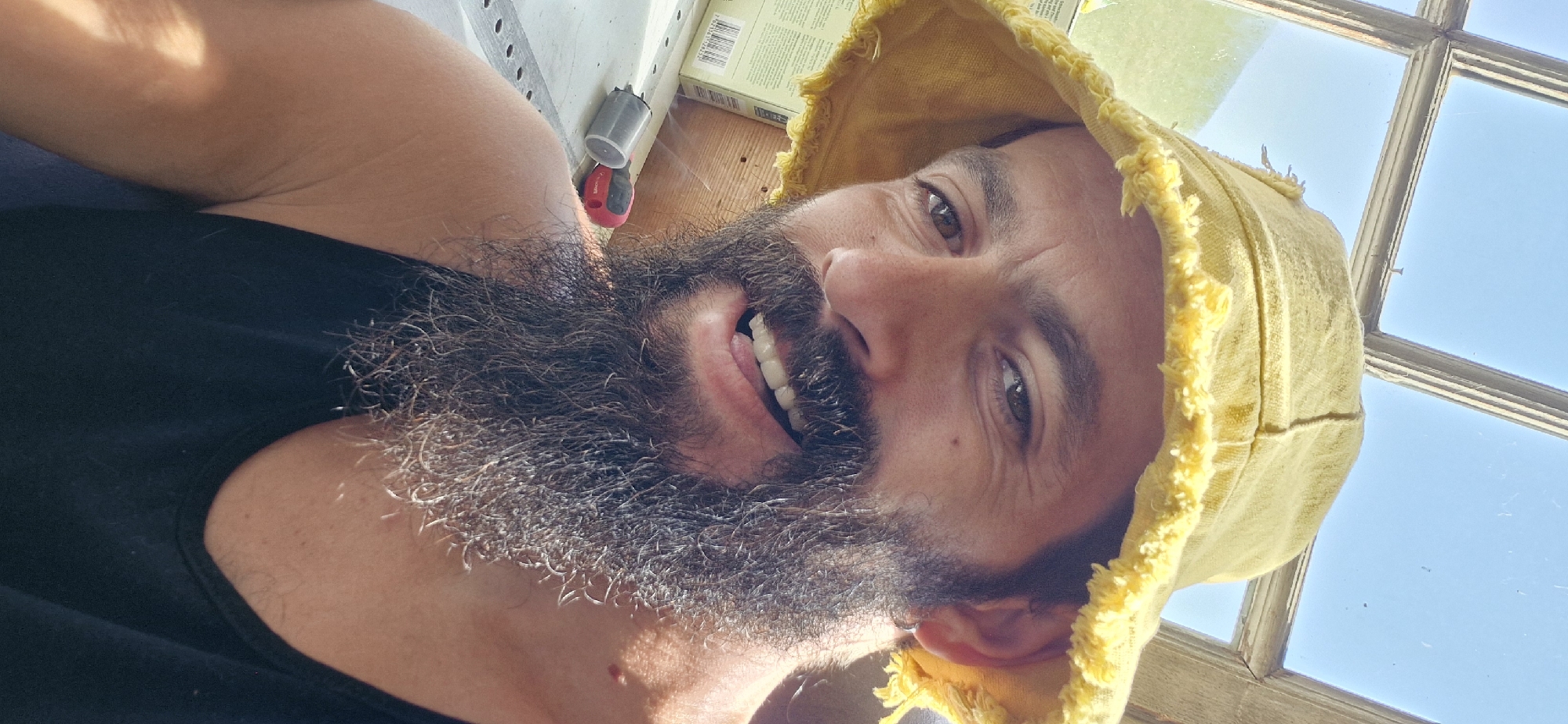 A man with a full beard and a yellow bucket hat is pictured in a selfie. He is wearing a black tank top and is looking at the camera with a slight smile. In the background, there is a window with a view of a bright blue sky and some wooden window frames. A few objects are visible on a surface in front of him, including what appears to be a tool and a cardboard box. The overall lighting suggests it is daytime. The image is cropped closely to the man's face and upper body, obscuring much of the surrounding environment. The scene is outdoors, possibly in a workshop or a shed, given the visible tools and materials. The man's relaxed posture and smile suggest he is in a comfortable and casual setting.