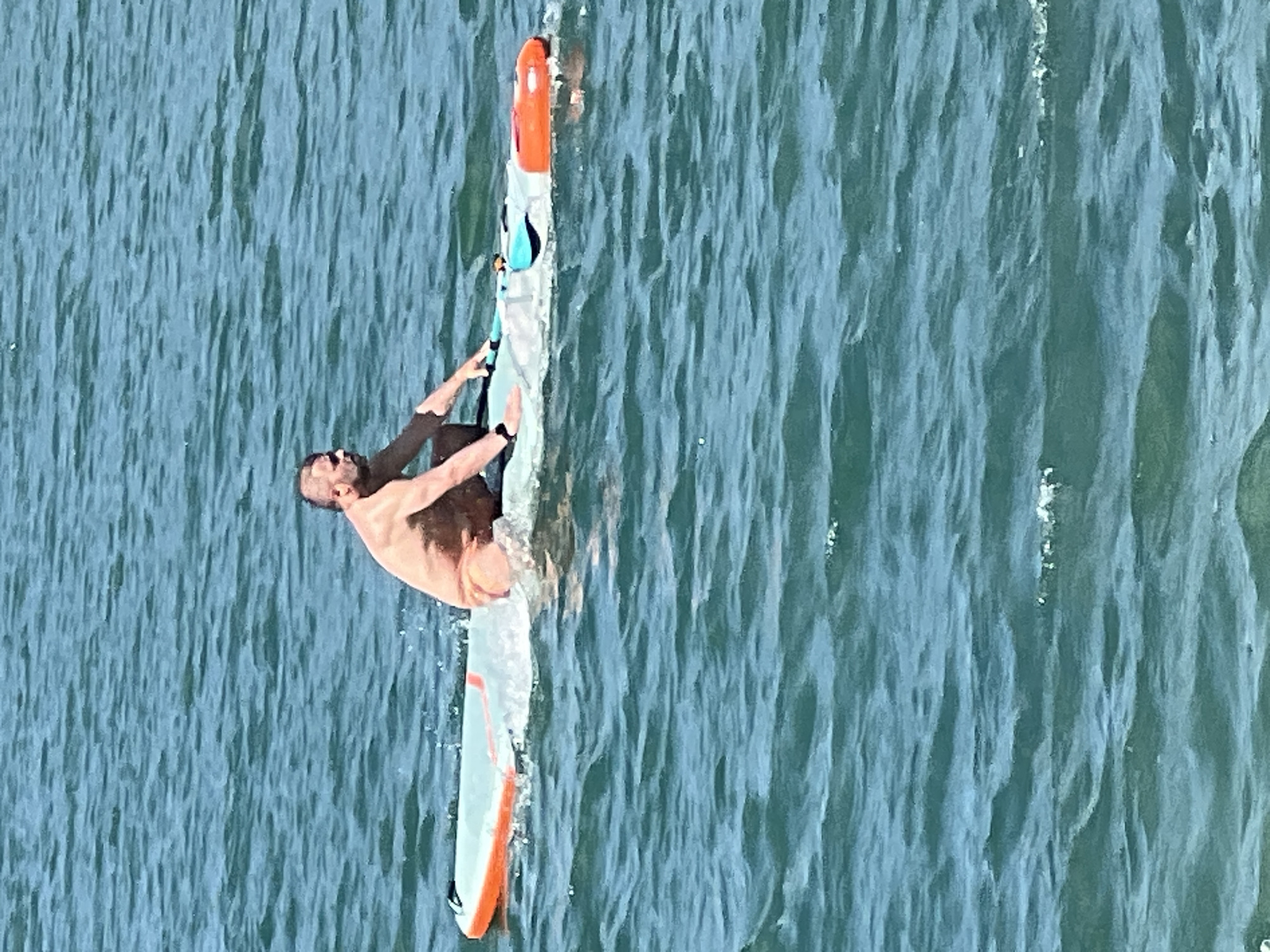 The image, taken from a high angle, shows a person windsurfing on a body of water. The individual, a bare-chested man with dark hair and a beard, is leaning over the windsurf board, seemingly in the middle of a maneuver. The windsurf board is long and slender, with orange and white detailing, and a dark blue and teal section near the front. The man is holding onto the boom with both hands, his body taut with effort. Splashes of water can be seen around the board, indicating movement.
The water is a deep, textured blue-green, with ripples suggesting a moderate choppiness. The lighting indicates it is daytime, likely sunny given the brightness on the water's surface. No specific landmarks are visible that would pinpoint the exact location, but the context provided places it in Vilnius, Lithuania. The activity is clearly windsurfing, a sport that combines elements of surfing and sailing. The man's posture and the dynamic water suggest a moment of action and skill. The image does not contain any visible text.