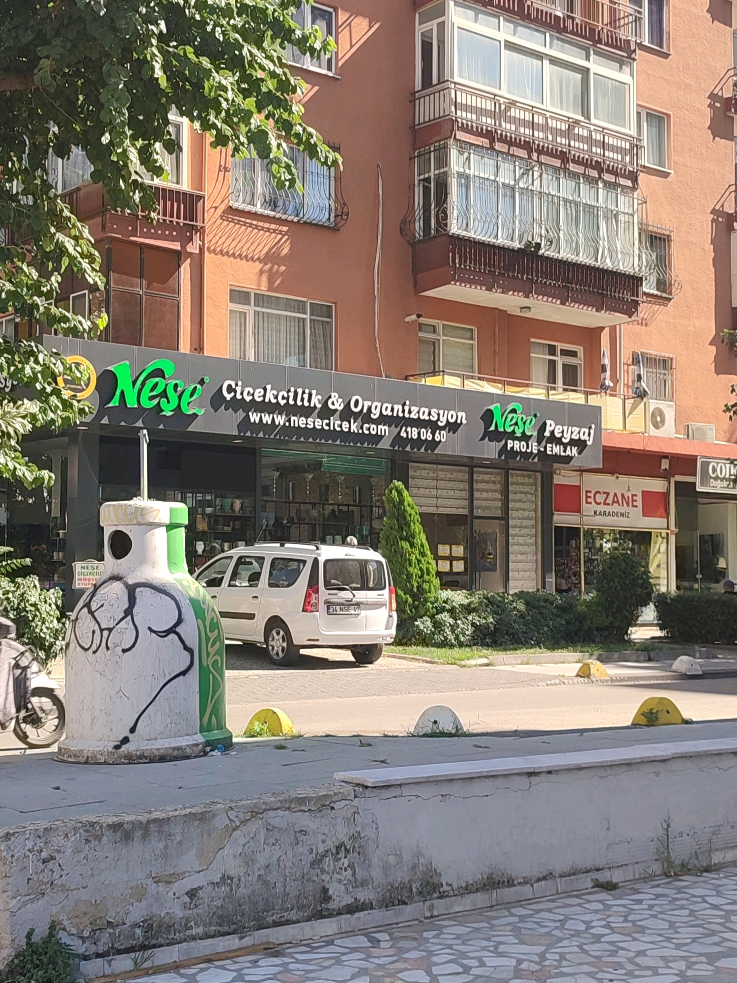 The image shows a street scene in Kadıköy, Turkey. In the foreground, a large, white, cylindrical waste bin with green trim and black graffiti art is visible on a paved sidewalk. A motorcycle is parked to the left of the bin, partially obscured by a tree's branches and leaves. Behind the bin, a white van with a license plate reading "34 NSE 40" is parked on the street.
A storefront with a dark gray facade and green neon signage is the central focus of the image. The sign reads "Nese Çiçekçilik & Organizasyon" and "www.nesecicek.com 418 06 60". To the right of this store, there is another storefront with a red and white sign that says "ECZANE KARADENIZ". Further to the right, a partial view of a store with a "CO" logo is visible.
The background consists of a building with multiple stories, featuring balconies with ornate railings and large windows. The building's facade is painted in a warm, peachy-orange hue. The sky is not visible in the frame. The environment appears to be an urban setting with shops and residential buildings. The time of day seems to be daytime, indicated by the bright sunlight casting shadows.