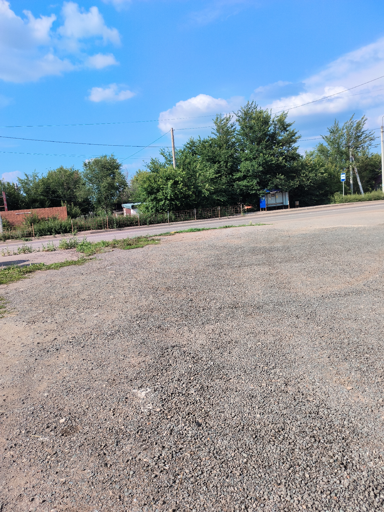The image captures a sunny day in Pokhvistnevo, Russia. The foreground is dominated by a gravel surface, appearing to be a roadside or parking area. Beyond this, a paved road runs horizontally across the mid-ground. On the opposite side of the road, a line of mature green trees forms a dense backdrop. Visible behind the trees are some structures, including a brick building on the far left and a bus stop shelter with a blue roof and side panels further to the right, identifiable by a blue bus stop sign. Power lines are strung across the sky, supported by utility poles, one of which is visible on the right side of the frame. The sky is a bright blue with scattered white clouds. The overall scene depicts an urban or semi-urban environment, possibly near a main thoroughfare or residential area.