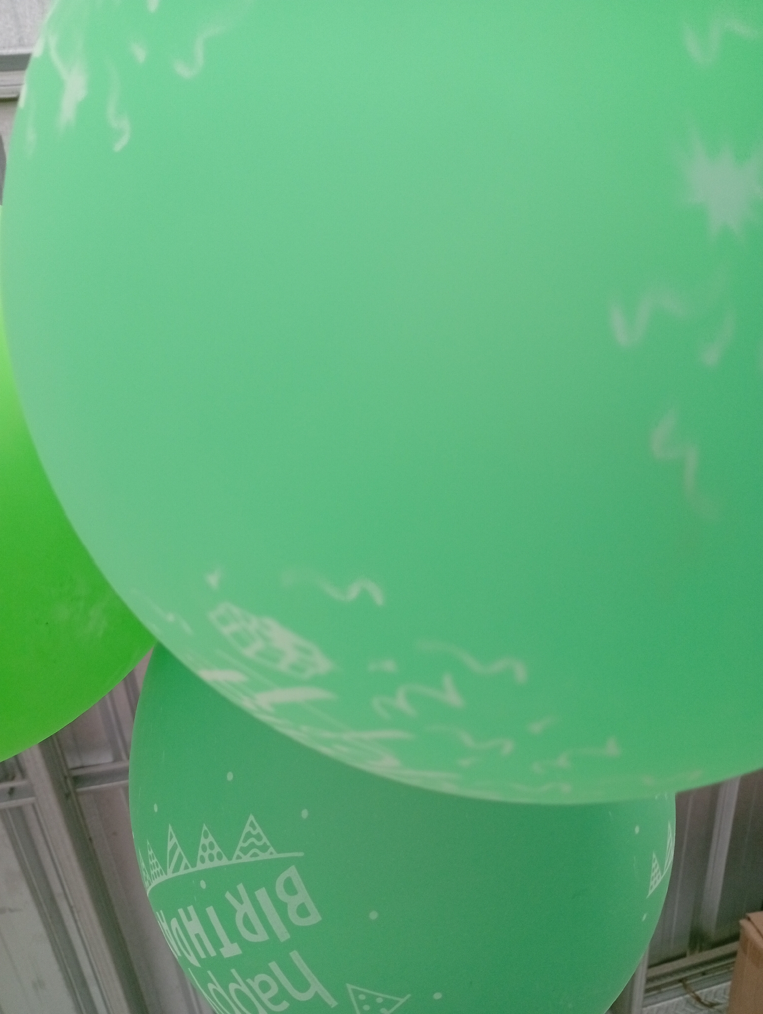 The image shows two green balloons, one larger and closer to the viewer, and another smaller one partially visible on the left. The larger balloon has white decorative patterns, including what appear to be party hats and dots, and text that reads "HAPPY BIRTHDAY" in reverse. The balloons are inflated and appear to be hanging or resting against a corrugated metal surface, possibly a ceiling or wall, which is visible in the background. The lighting suggests an indoor or shaded outdoor environment. The scene is in Balung, Indonesia.