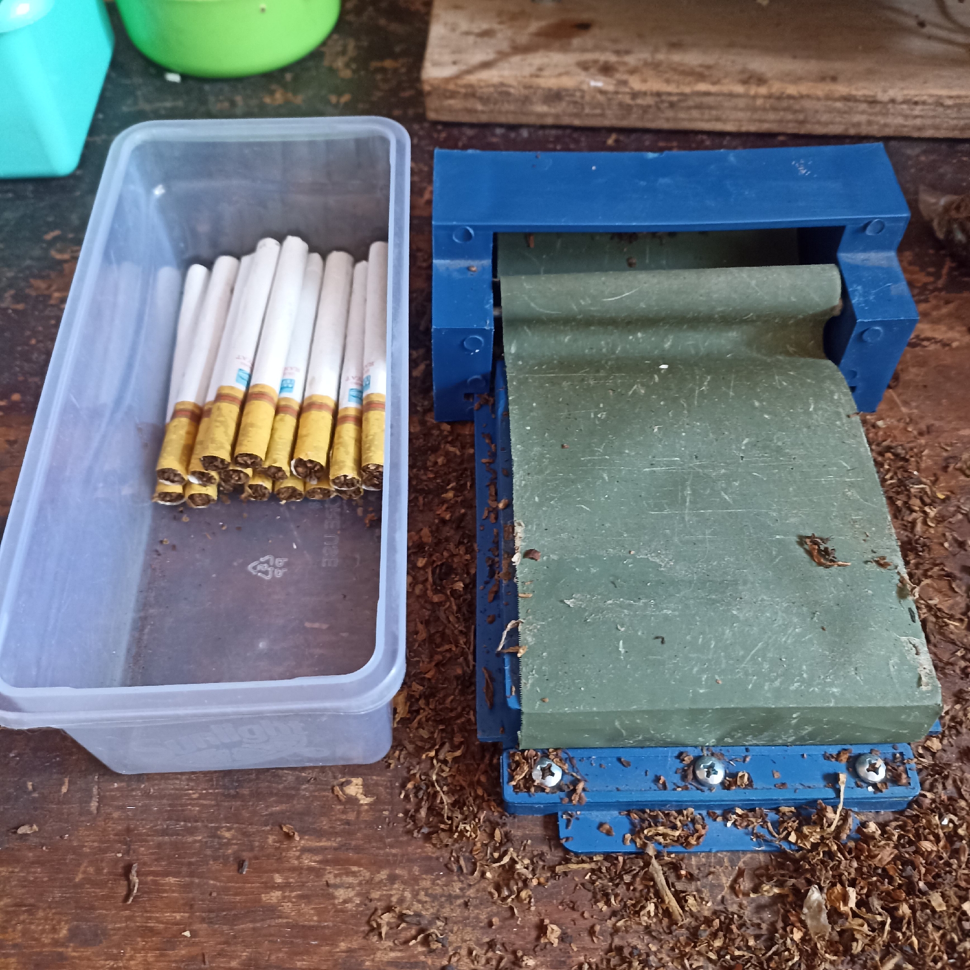 The image shows a close-up, overhead view of a manual cigarette rolling machine on a wooden surface. To the left of the machine, a clear plastic container holds several unrolled cigarettes. The cigarettes are white with yellow bands near the filter ends, which are filled with tobacco. Scattered around the cigarette rolling machine and on the wooden surface are loose tobacco leaves. The cigarette rolling machine itself is blue and green. The wooden surface appears to be aged and somewhat worn. The overall scene suggests a process of hand-rolling cigarettes. The specific location in Magelang, Indonesia, is not indicated by any visual cues within the image.