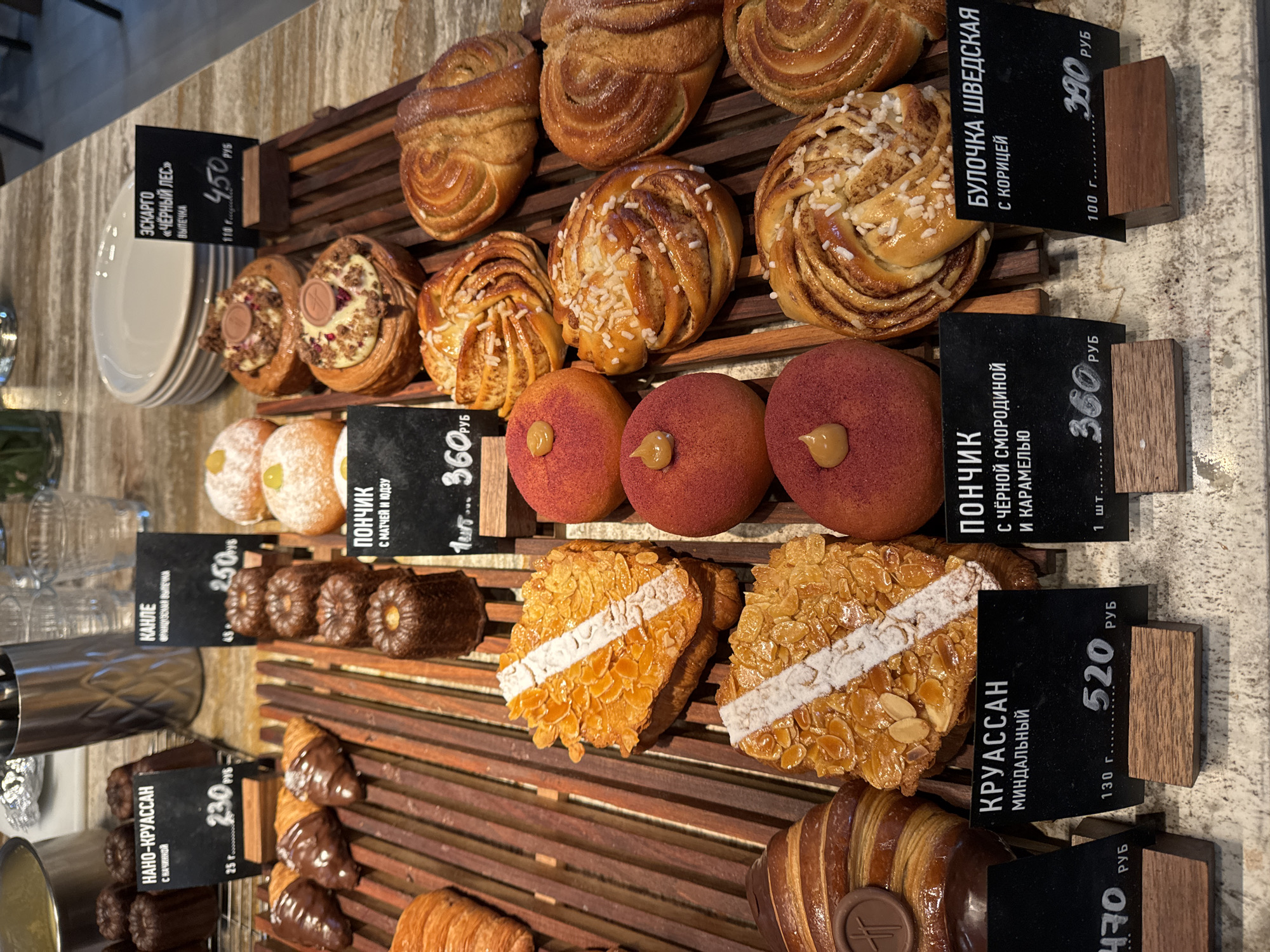 A display of baked goods in a store, likely a bakery or cafe. The baked goods are arranged on wooden slats. The baked goods consist of: pastries, croissants, canelés, donuts and Swedish cinnamon rolls. Each item has a small sign with its name and price in Russian rubles. Some items present are “Éscargot” Black Forest, “Nano-croissant” filled, Canelés, Donuts, and Swedish cinnamon rolls. The image is taken at eye level. The location appears to be in Unknown, Unknown.