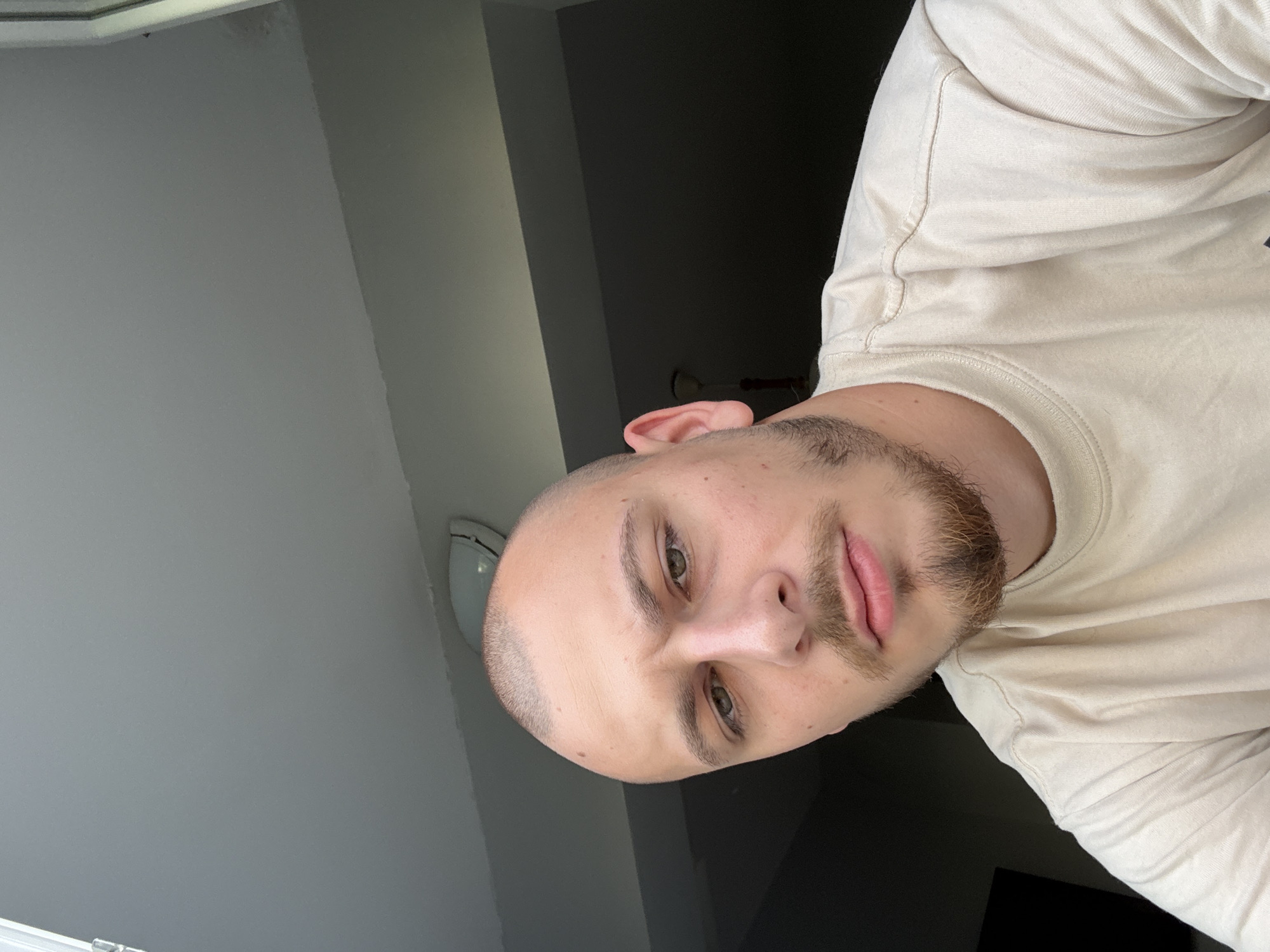 The image is a close-up selfie of a bald man with a light beard and mustache. He is wearing a light-colored t-shirt. The background appears to be indoors, with a ceiling visible above him. The lighting suggests it is daytime. No specific details can be determined about the location within the city of Stara Zagora, Bulgaria from the provided image.