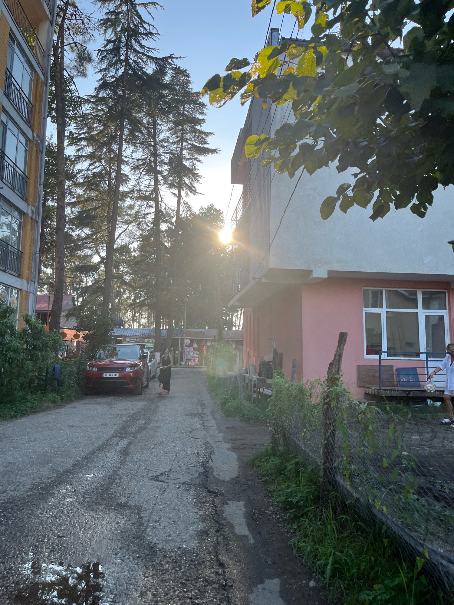 The image depicts a street scene in Ureki, Georgia, likely in the late afternoon given the low angle of the sun. On the left side of the image, a multi-story building with balconies and windows is visible, constructed with yellow and grey materials. Tall evergreen trees are interspersed along the street and behind the buildings, with sunlight filtering through their branches.
The street itself is paved with asphalt, showing signs of wear and puddles from recent moisture. A red SUV is parked on the left side of the street. A woman in a dark dress is walking away from the camera down the street, while another person, partially visible in a white top, is standing near a building on the right side of the image.
On the right side, a pink-colored building with a light grey upper section is prominent. It features large windows and a small porch area. A wire fence covered in vegetation runs along the foreground of this building. Small stalls or shops are visible in the background, suggesting a commercial area. The overall impression is a somewhat rustic street in a resort town setting.