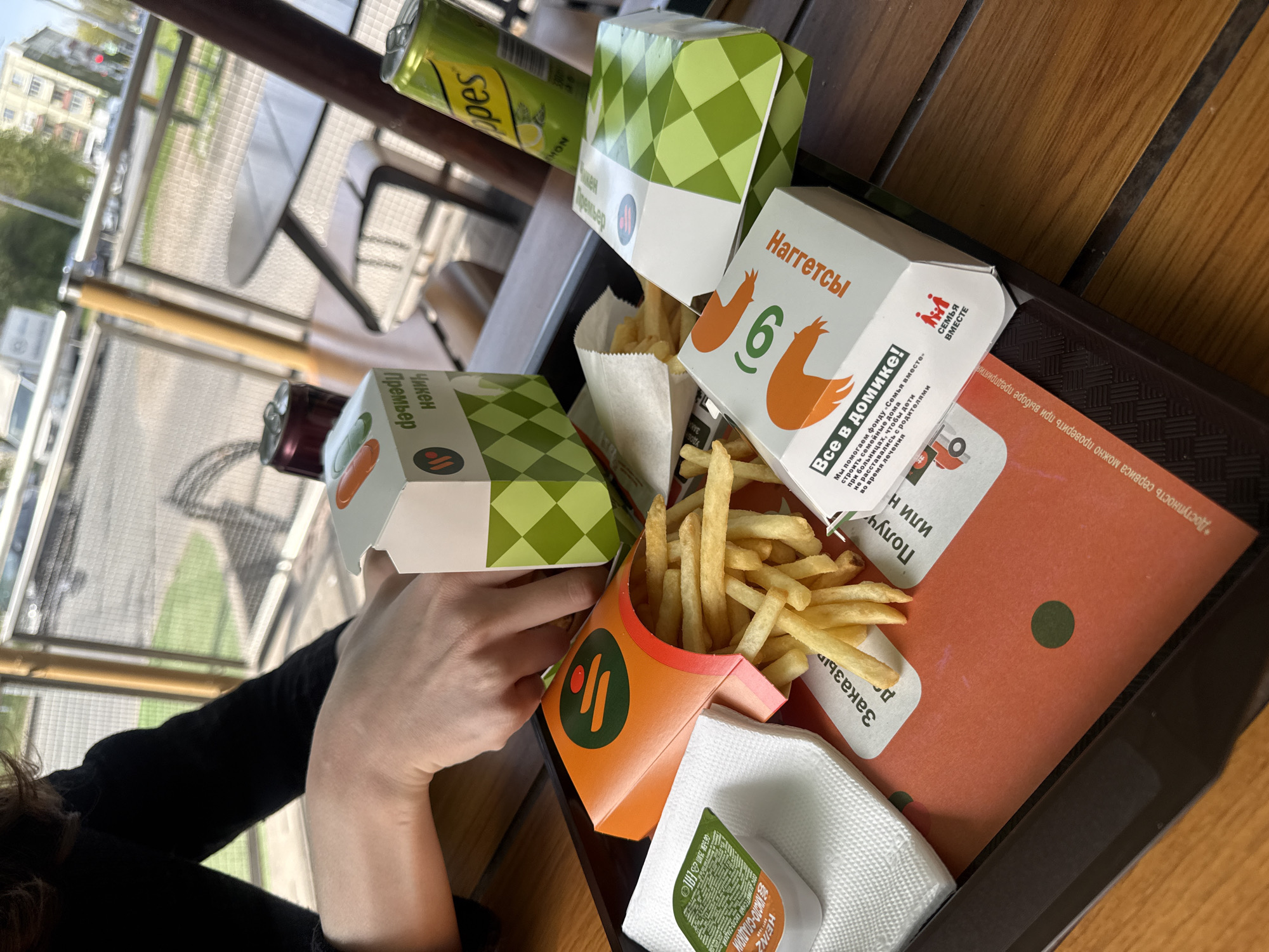Here is a detailed description of the image:
The image shows a tray of fast food items on a wooden table. A person's hand is reaching towards a "Chicken Premier" box, which is green and white with a diamond pattern. Other items on the tray include a "Nuggets" box, a container of French fries in an orange and black branded box, a small container of Heinz sauce, napkins, and a Schweppes lemon drink. The "Nuggets" box indicates that there are six nuggets. There's also a promotional paper mat beneath the food items, with a dark brown tray beneath. Outside, chairs and tables are visible through a window, suggesting a cafe or restaurant setting. Text is visible in Russian on several of the containers. The location is likely Saint Petersburg, Russia.
