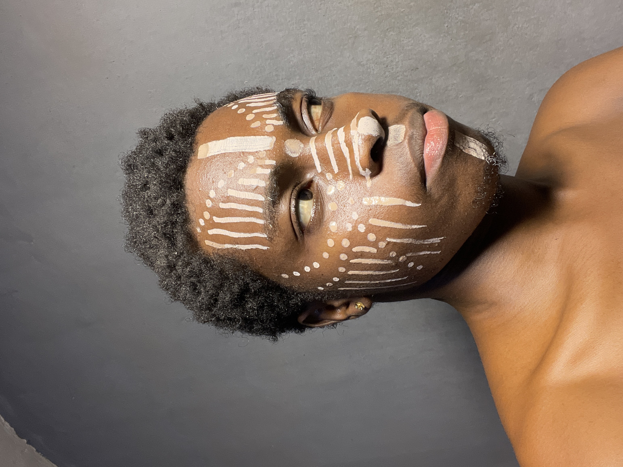 A close-up shot presents a dark-skinned person with short, tightly curled dark hair. Their face is adorned with tribal-style markings in a light cream color, consisting of horizontal lines and dots. The person is slightly angled, with their head tilted upwards and their gaze directed off-frame. A small gold earring is visible in their left ear. The background is a solid, neutral gray. The scene's location in the city of Nkpor, Nigeria is not discernible.