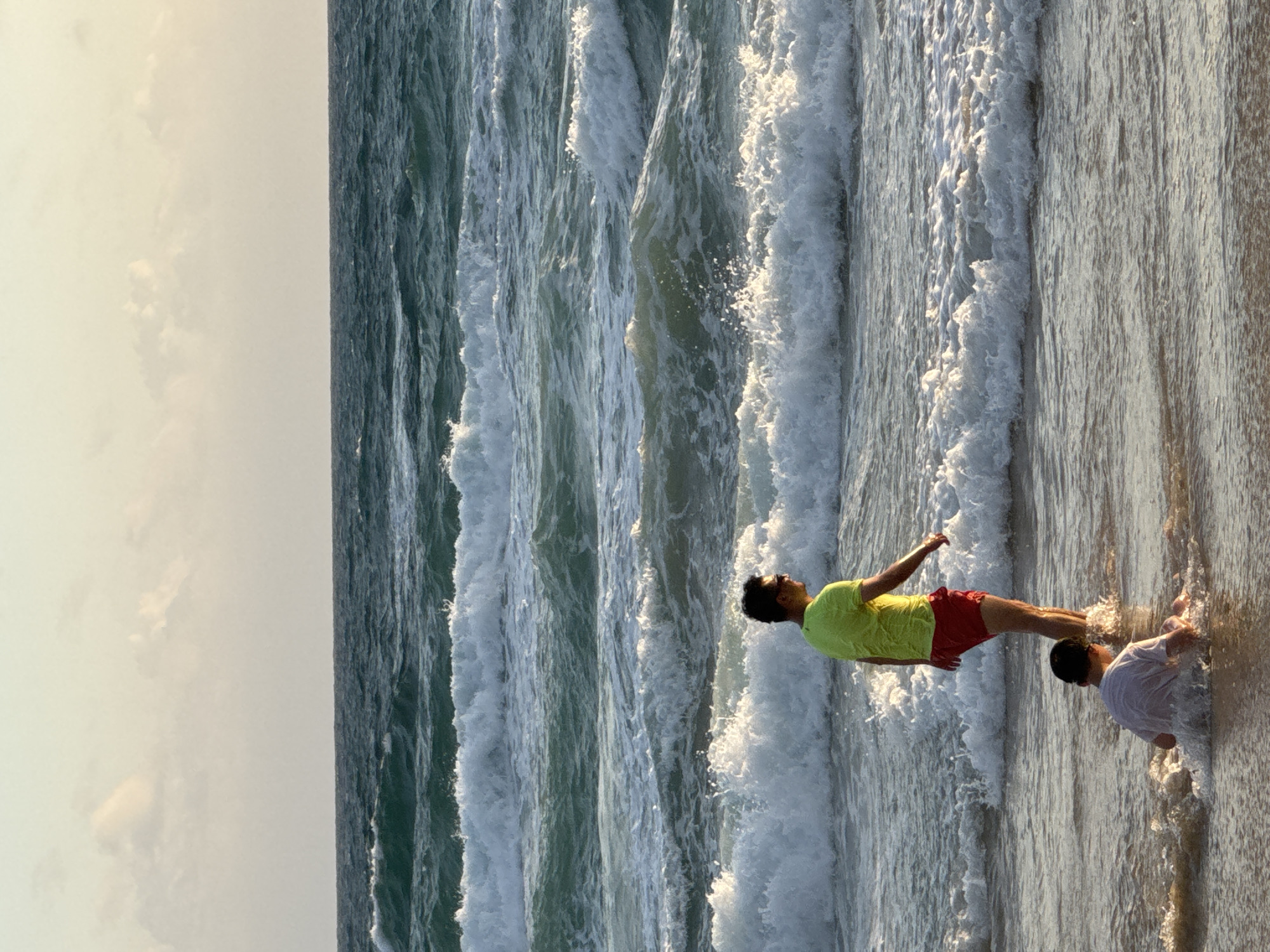 A man and a child are in the shallow water of Karon Beach in Thailand. The man, wearing a bright green t-shirt and red shorts, stands facing away from the camera, looking out at the ocean. The child, wearing a white t-shirt, sits in the water near the shore. The ocean is dark blue and choppy, with several waves breaking in the foreground and background. The sky is pale with a few scattered clouds. The lighting suggests it is late afternoon or early evening.