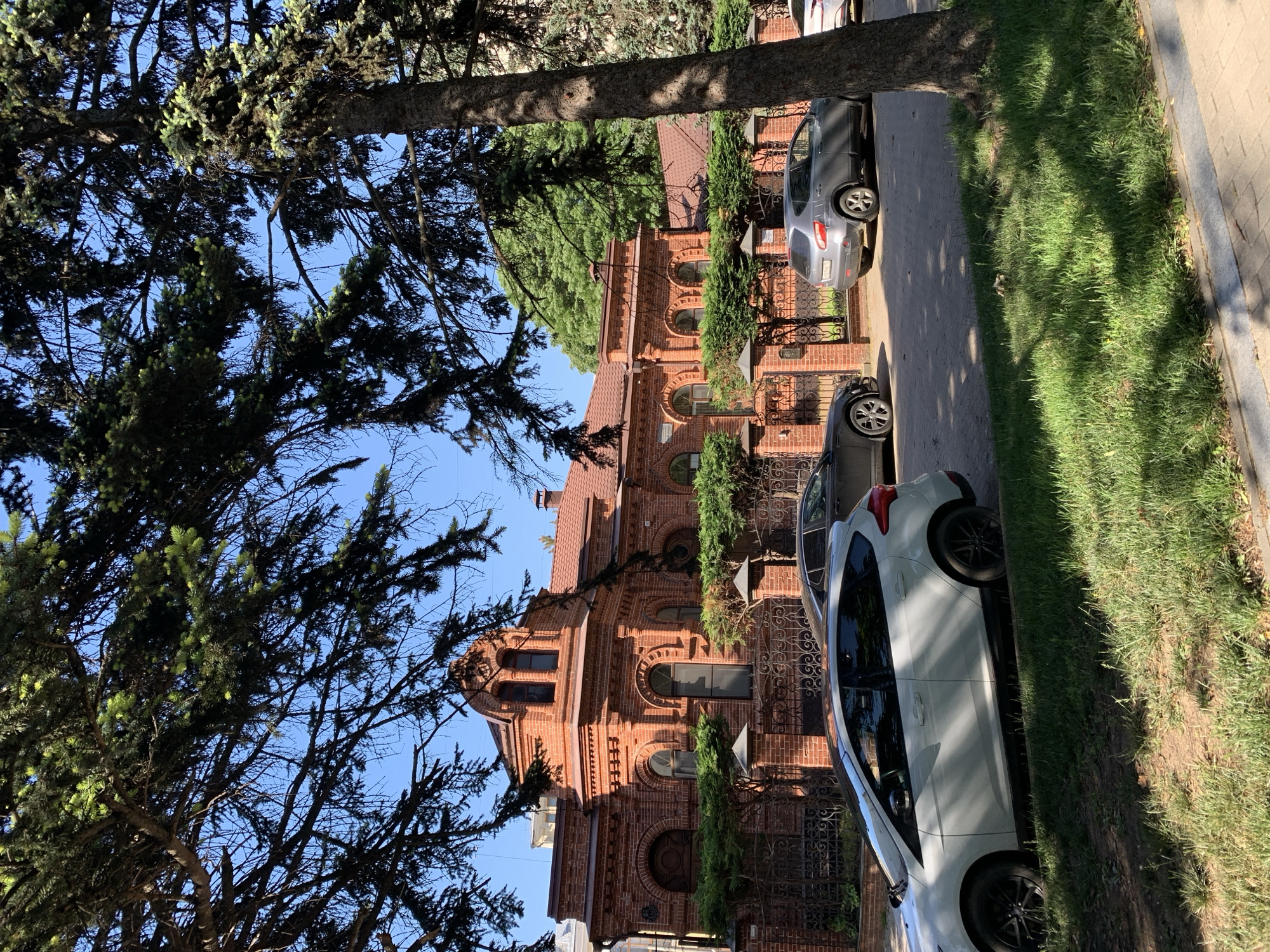 Here is a description of the image:
The image depicts a street scene in a city. The location appears residential, with a prominent brick building visible in the background. The building features architectural details, including arched windows and a decorative roof. Several cars are parked along the side of the street. A tall tree stands on the left side of the frame, partially obscuring the view with its branches. A sidewalk and grassy area are visible on the right side of the image. The weather seems sunny and clear.