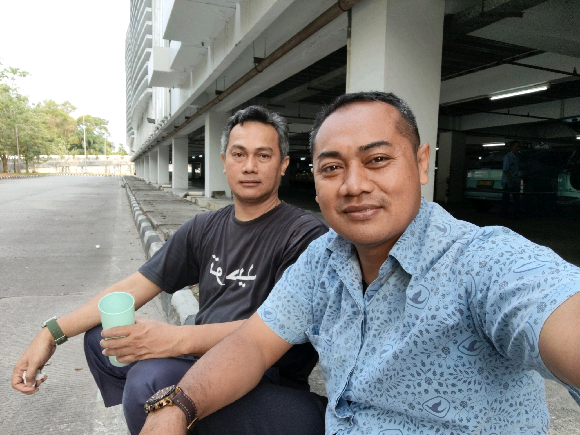 Two men are seated next to each other outdoors, with a large modern building in the background. The man on the left has short, dark hair with some graying at the temples and is wearing a black t-shirt with white Arabic-style lettering and dark pants. He is holding a light green plastic cup in his left hand and has a green watch on his left wrist. The man on the right has short, dark hair and is wearing a light blue patterned short-sleeved button-down shirt and dark pants. He has a brown and gold watch on his right wrist. They are both looking towards the camera. The background consists of a multi-story building with many balconies and a parking structure. The ground is paved with asphalt. The scene appears to be in a parking lot or outdoor public space in Balikpapan, Indonesia, given the architectural style and vegetation visible.