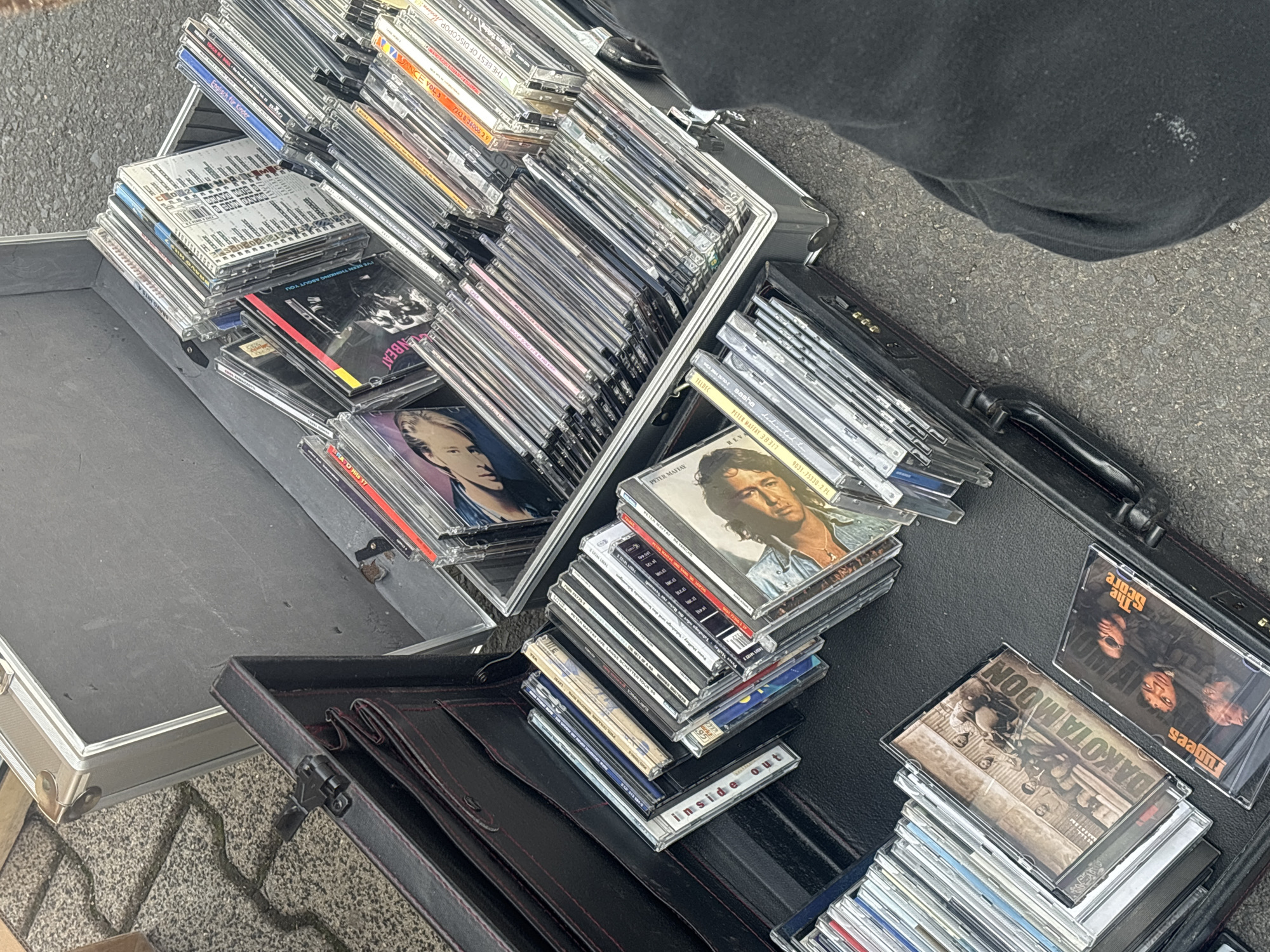 The image depicts an assortment of compact discs (CDs) arranged in open briefcases, likely at a street sale or flea market in Eichenzell, Germany. The CDs are stacked haphazardly, revealing a variety of album covers. The surrounding environment appears to be an outdoor setting with gray pavement or asphalt visible. The overall scene suggests a commercial activity involving the sale of used or vintage music.