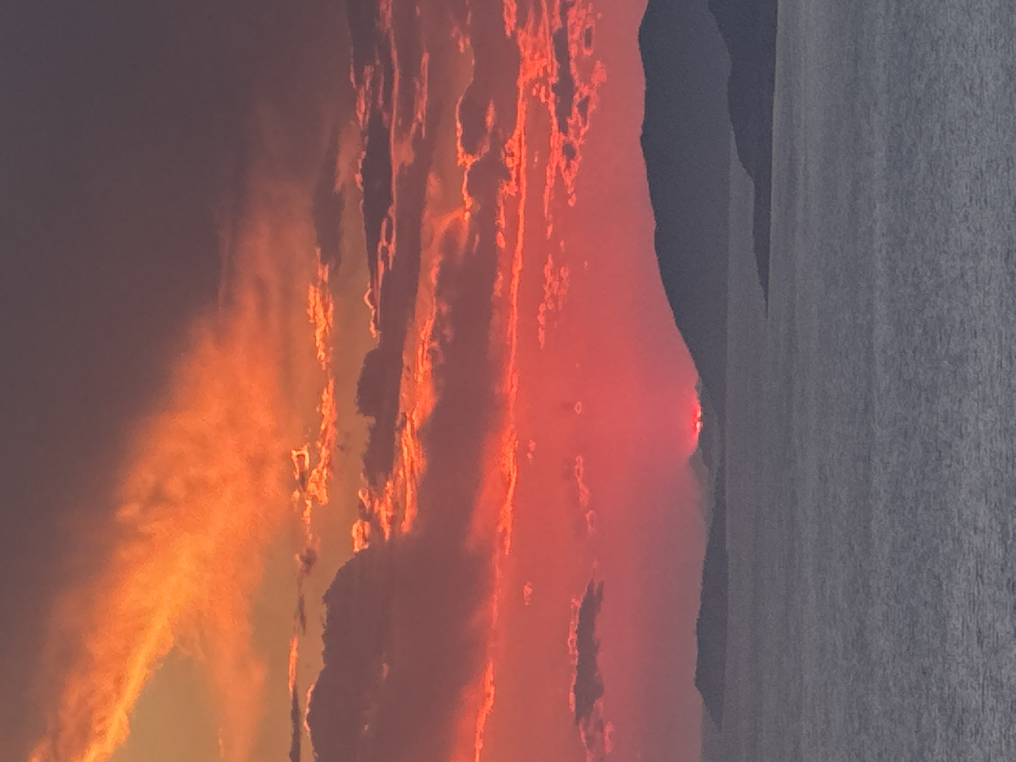 An expansive, dramatic sunset paints the sky in fiery hues of orange and red over the Adriatic Sea. Silhouetted islands with gentle slopes jut out from the water, their dark forms stark against the vibrant sky. The water itself is a deep, placid grey, reflecting the subdued light of dusk. The sun, a vivid crimson orb, is partially obscured by the horizon, casting a soft glow on the surrounding clouds. The scene suggests a serene and breathtaking moment captured from a vantage point overlooking the Montenegrin coast near Zagora.