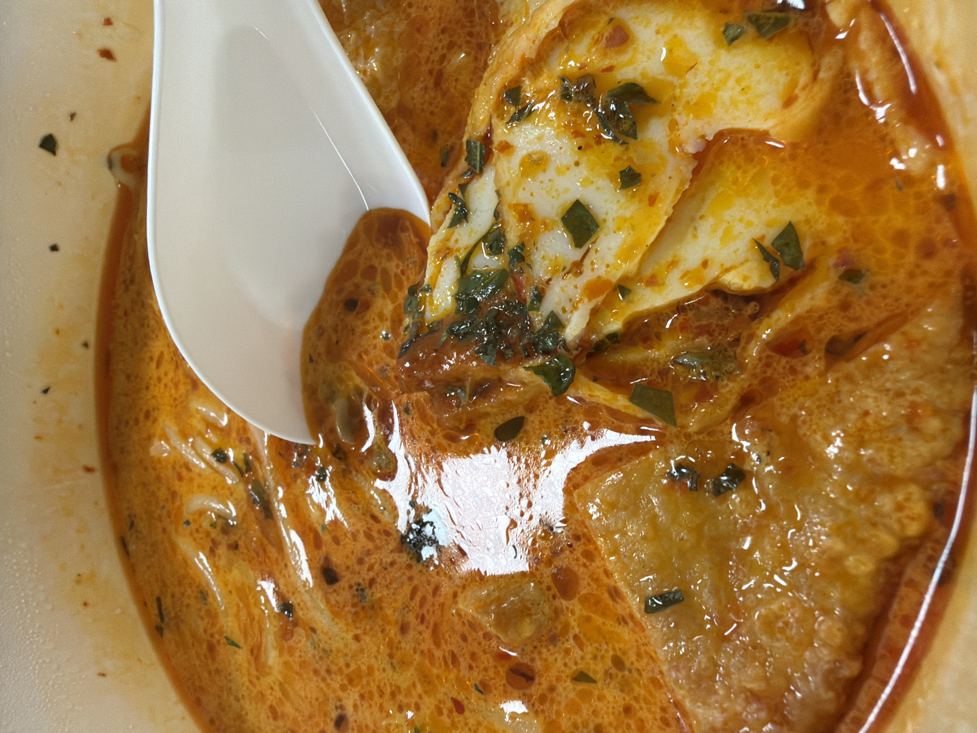 A close-up overhead shot shows a bowl of curry laksa, a popular Singaporean dish. A white spoon is partially submerged in the rich, orange broth, which is topped with pieces of white fish, tofu, and scattered green herbs. The bowl appears to be made of paper or a similar disposable material, suggesting a takeout or casual dining setting. The lighting is bright and even, highlighting the textures and colors of the food. The background is out of focus, but hints at a simple, possibly indoor environment.