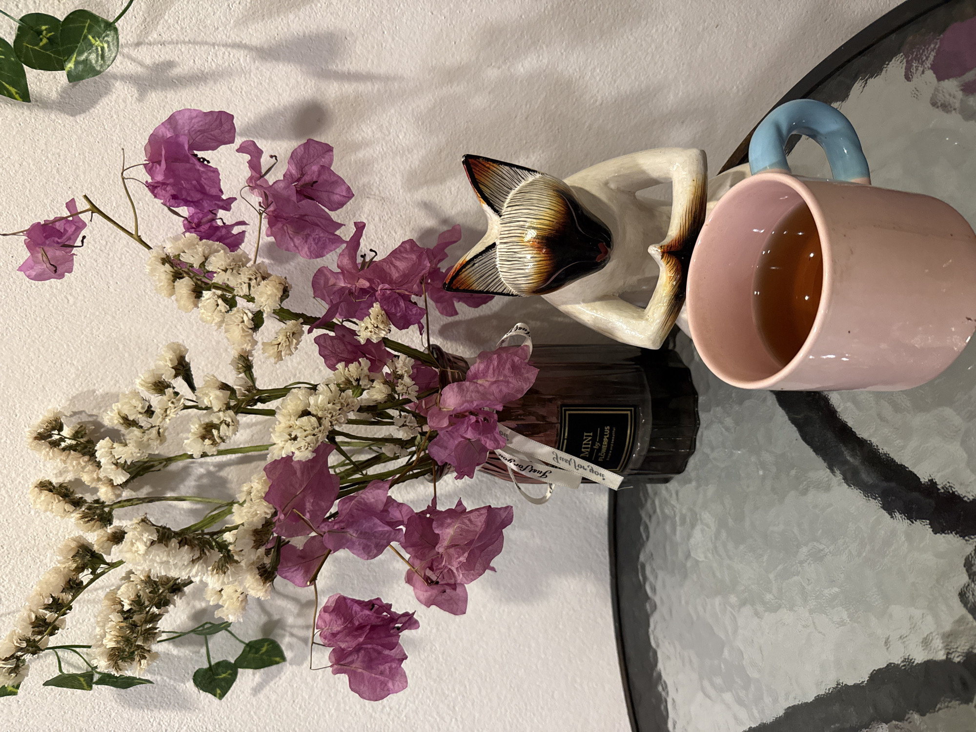 An indoor still life photograph shows a glass-top table with several objects arranged on it. In the background, a white wall displays shadows cast by objects not fully visible. A bouquet of dried white flowers and purple bougainvillea blooms is arranged in a dark glass vase with a ribbon and a small black label that reads "MINI by flowerplus Simple and beautiful." To the right of the vase sits a wooden figurine of a cat in a meditative pose, its face painted with black, white, and orange details, and pointed ears. In the foreground, a pink mug with a light blue handle, filled with a brown liquid, rests on the table. The table surface has a reflective, textured pattern. The overall scene depicts a tranquil, decorative setting. This scene is not specific to Ko Samui District, Thailand and could be found in any location.