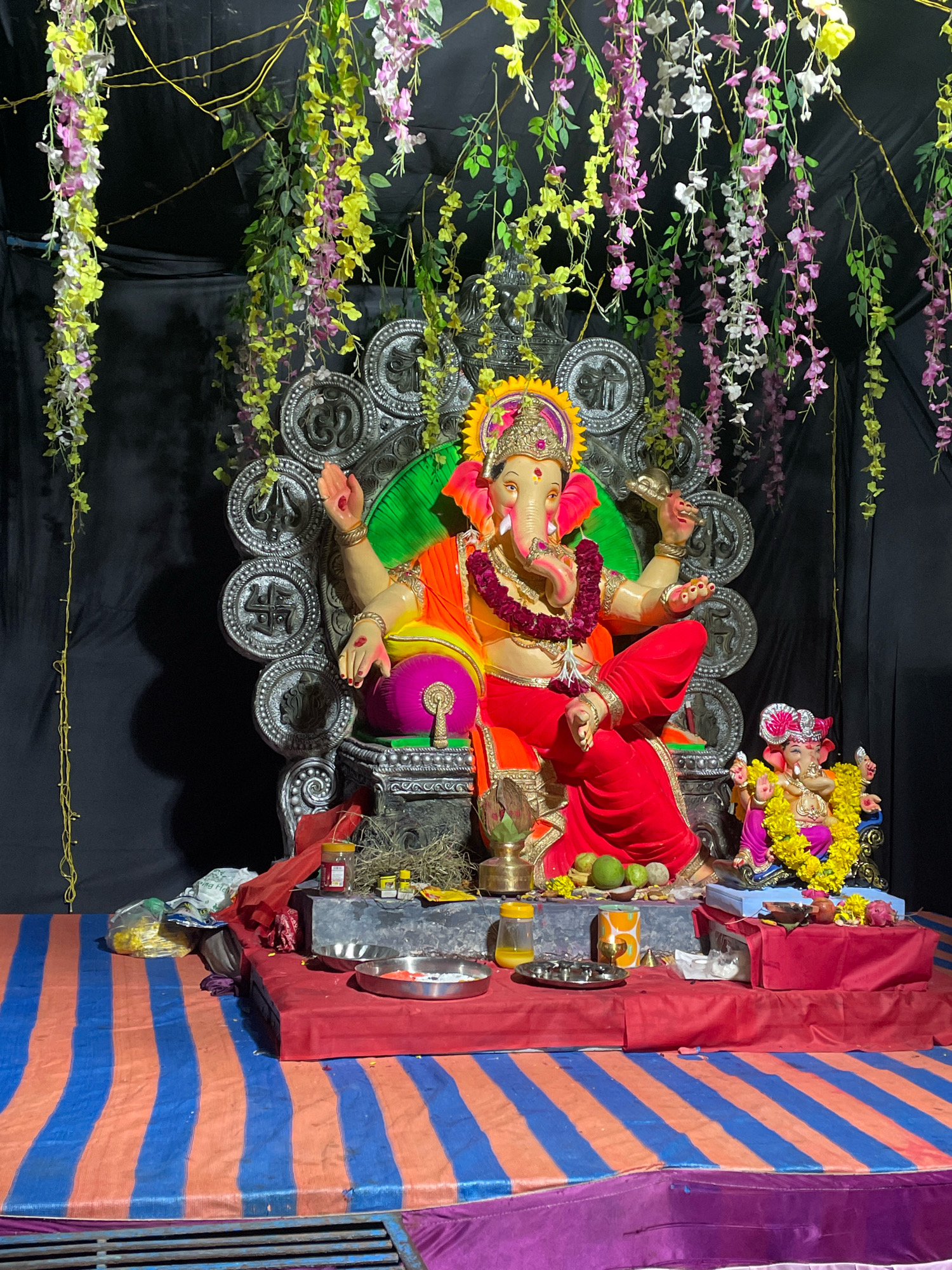 In Ahmedabad, India, a large, elaborately decorated statue of the deity Ganesha is the central focus of this scene. The statue, depicted with multiple arms and wearing vibrant orange and red attire, is seated on an ornate, silver throne. A smaller Ganesha statue, adorned with yellow flowers, is positioned to the right of the larger one. The statues are placed on a raised platform covered with a red cloth, in front of which a blue and orange striped cloth serves as a ground covering. Various offerings, including fruits and what appear to be small containers, are visible on the platform. Cascading artificial flowers in shades of yellow, pink, and white hang from above, creating a festive backdrop against a black curtain. The overall presentation suggests a religious or cultural celebration, likely Ganesh Chaturthi, taking place in Ahmedabad.