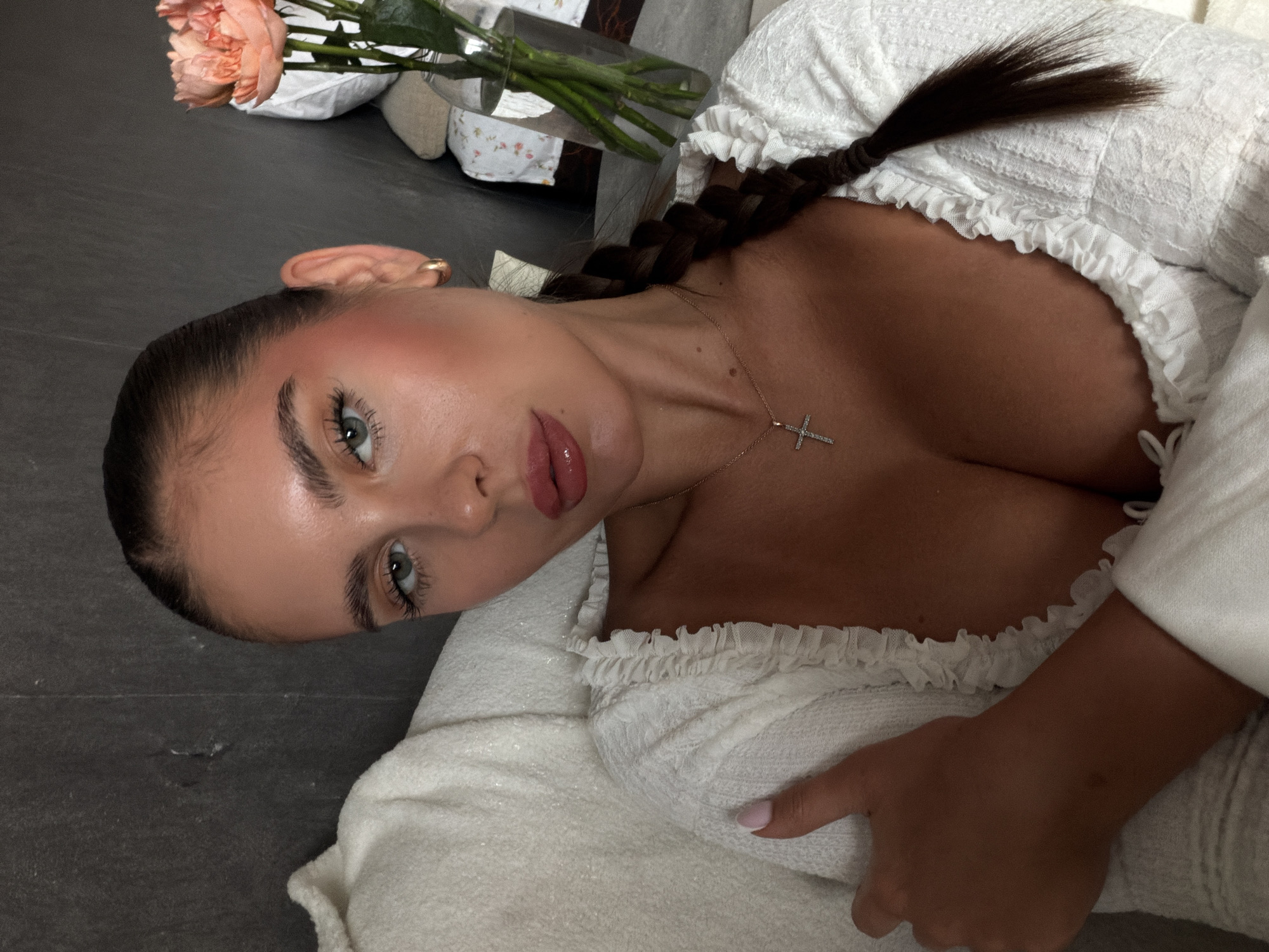 A woman lies on a light-colored, textured surface wearing a white, ruffled top and a silver cross necklace. Her dark hair is braided. Her head is turned towards the camera. A vase with pink flowers is visible in the background. The location is not discernible as Krasnoyarsk, Russia based on the provided visual information.