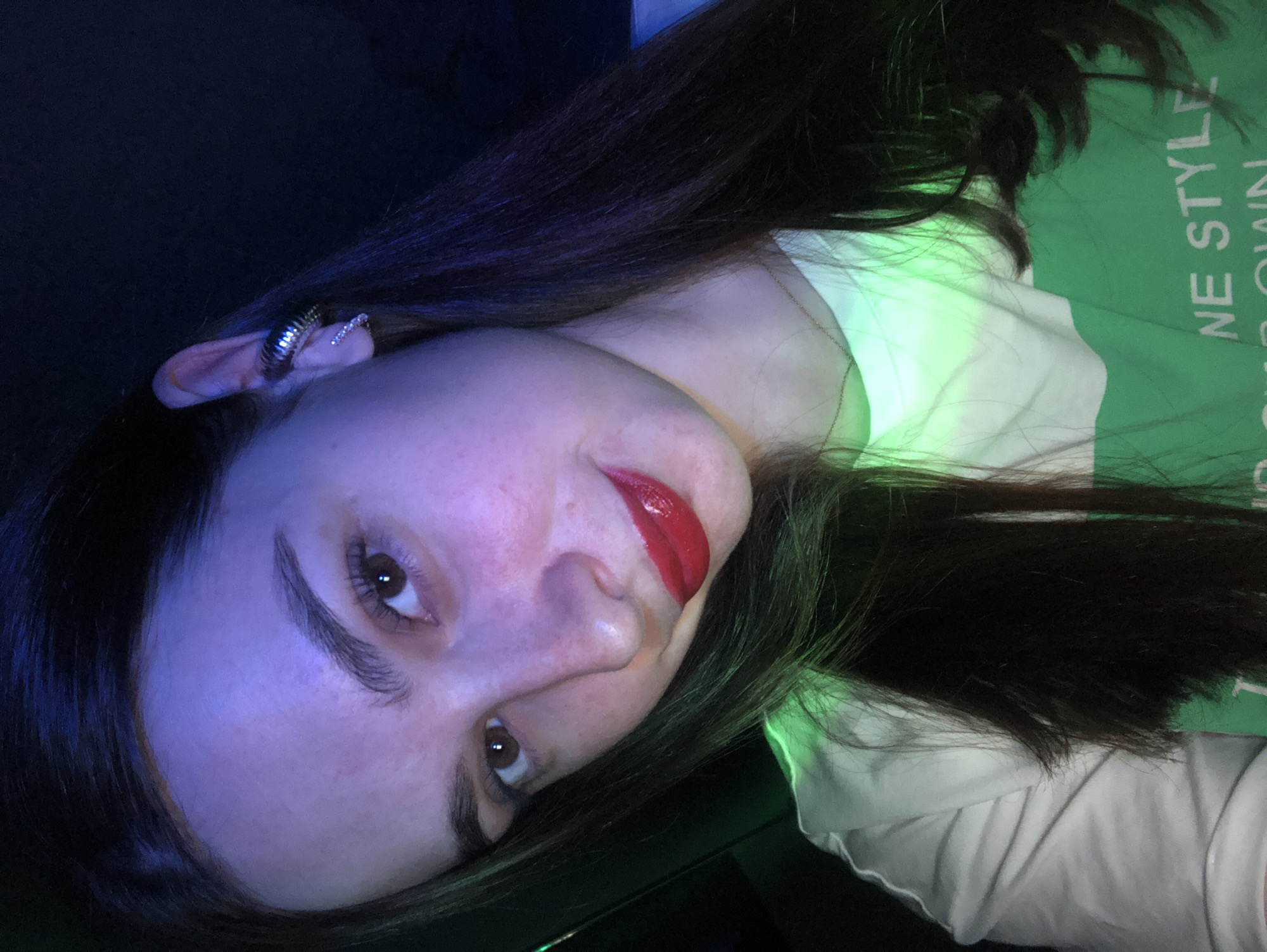 A woman is seen at an unusual angle in a dimly lit setting. Her face is slightly angled upwards, illuminated by blue and green light. She has dark brown hair, red lipstick, and several earrings in her left ear. She appears to be wearing a white shirt with green lettering that reads "NE STYLE UR OWN". The background is dark and indistinct. There is no location in the city of Noviy, Russia.