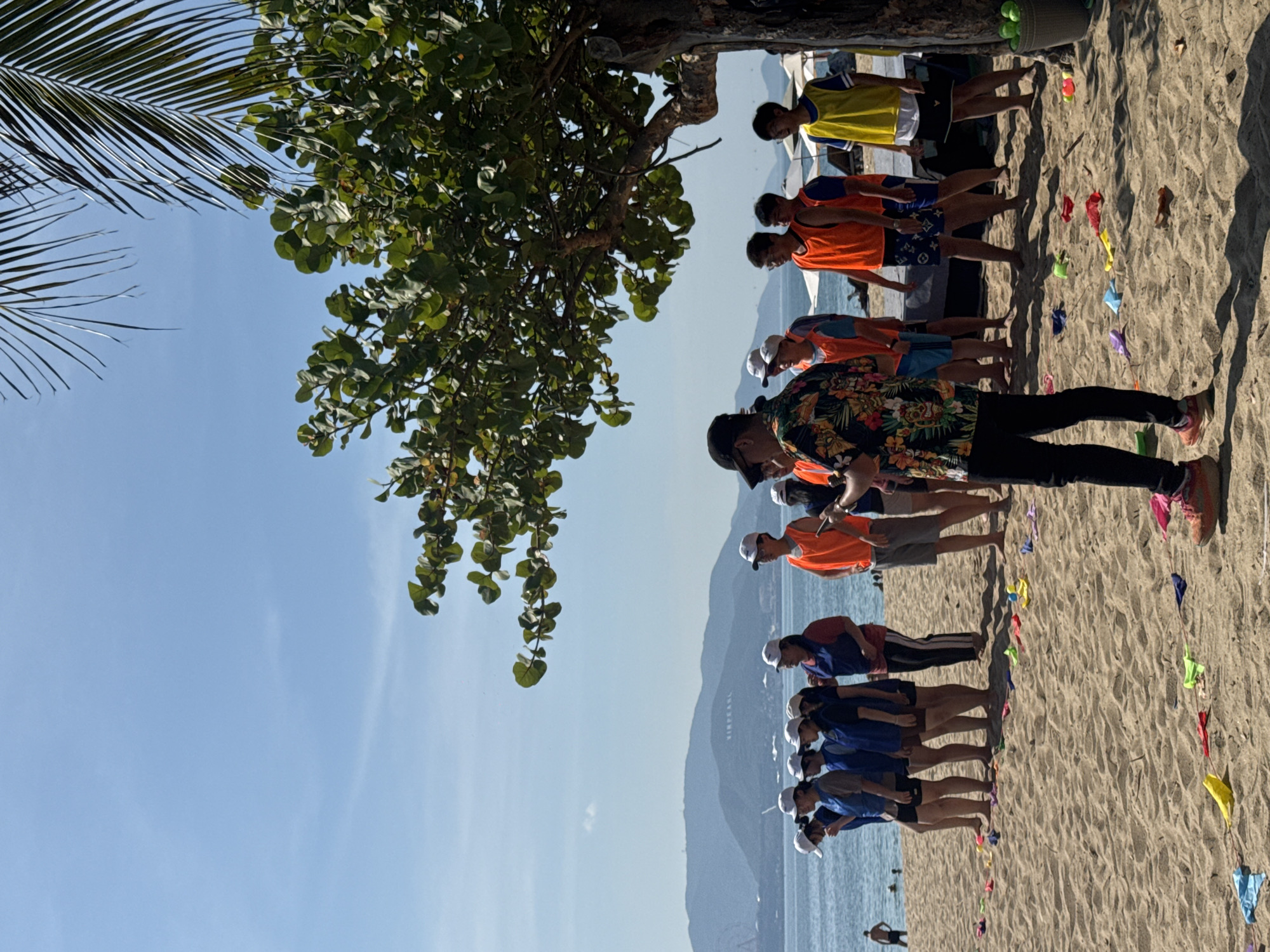 A group of approximately 15 people, some wearing orange and yellow vests, are gathered on a sandy beach. They appear to be participating in an outdoor activity or team-building exercise. Several individuals are wearing white caps. In the background, the ocean is visible, with mountains and a Ferris wheel in the distance, suggesting the location is Nha Trang Ward, Vietnam. The sand is marked with colorful items, possibly part of a game. A person in the foreground is wearing a floral shirt and dark pants, and another person to their right is wearing an orange vest and grey shorts. A large tree with green foliage shades part of the scene. The sky is clear and blue.