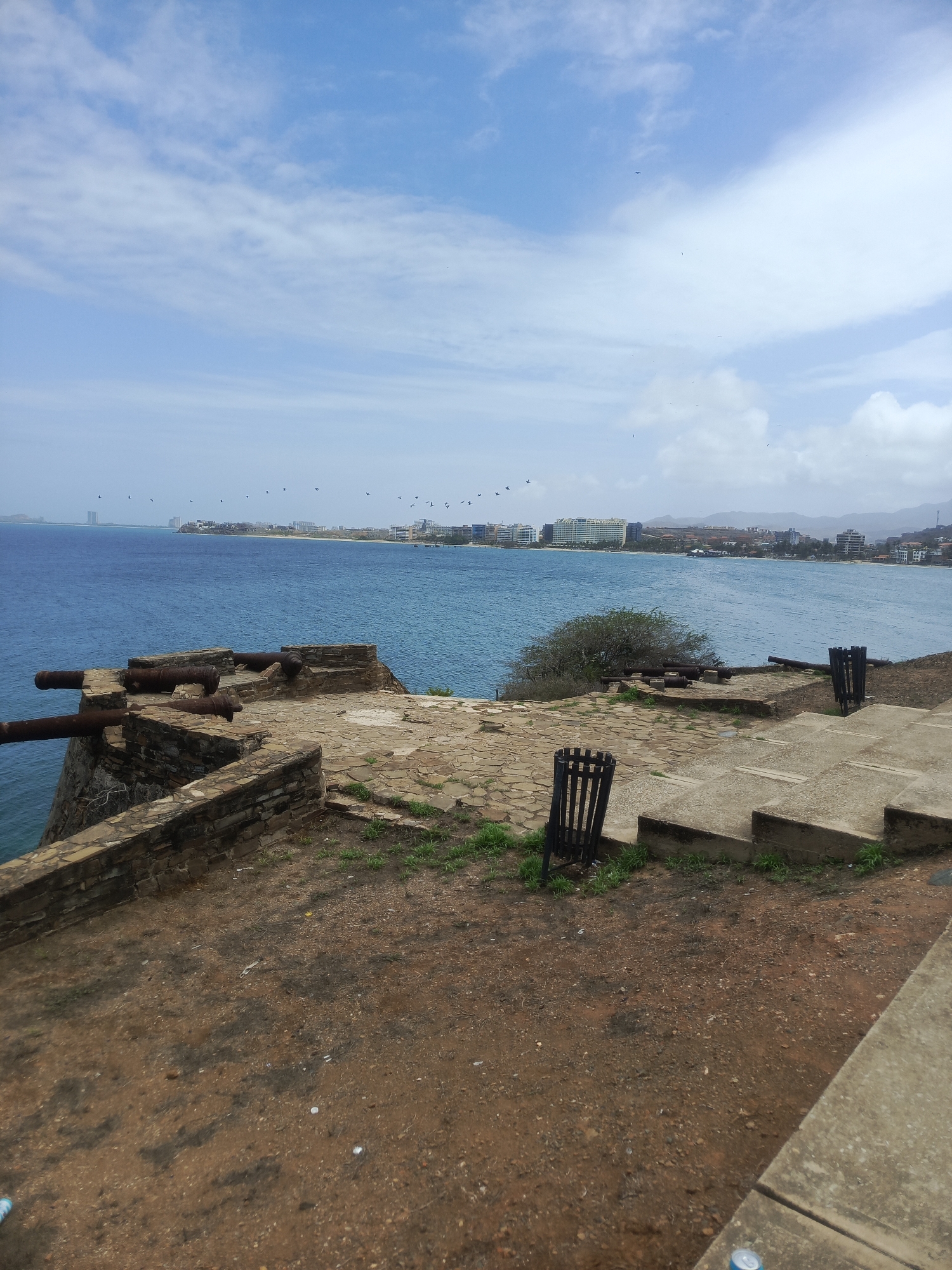 A wide-angle, eye-level outdoor shot captures a view of the Caribbean Sea from an elevated vantage point overlooking the city of Pampatar, Venezuela. In the foreground, a weathered stone fortification with several antique cannons is situated on a dirt and stone-paved area. Two black metal trash receptacles are visible on the right side of the frame, near concrete steps leading down. Beyond the fortification, the calm, blue sea stretches out to the horizon, where the city's skyline is dotted with buildings of varying heights. The sky above is a pale blue, with scattered white clouds and numerous small birds in flight. The scene appears to be a historical landmark offering a scenic view of the coastal city and its surrounding waters.
