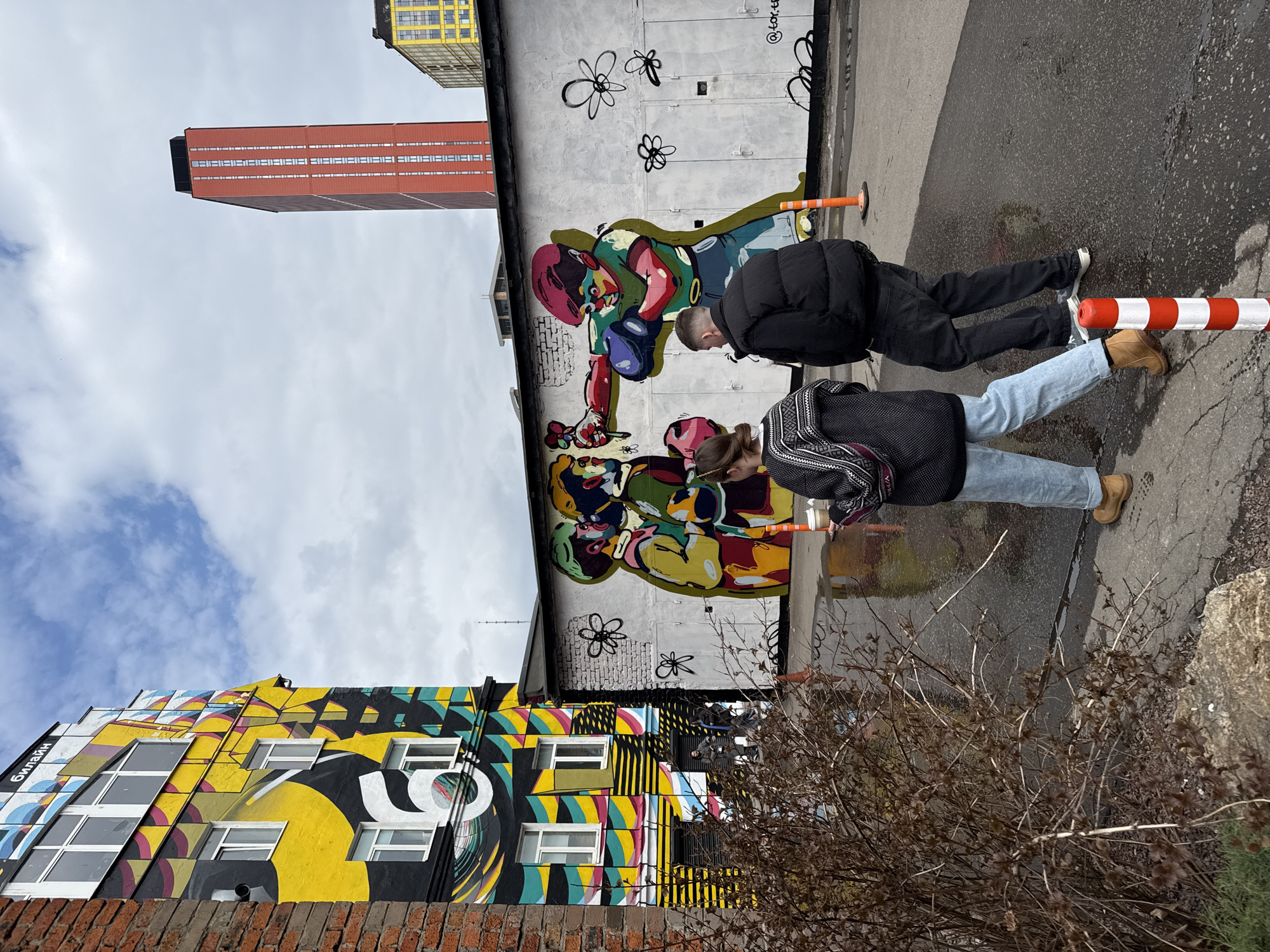 The photograph was taken outside in an urban environment, and shows two pedestrians standing in front of a concrete building with a mural of two colorful figures. To the left of the concrete building is a brick building with a multi-colored abstract design. Tall buildings can be seen above both buildings. A dry, brownish bush is in the lower right of the image. The location is Moscow, Russia.