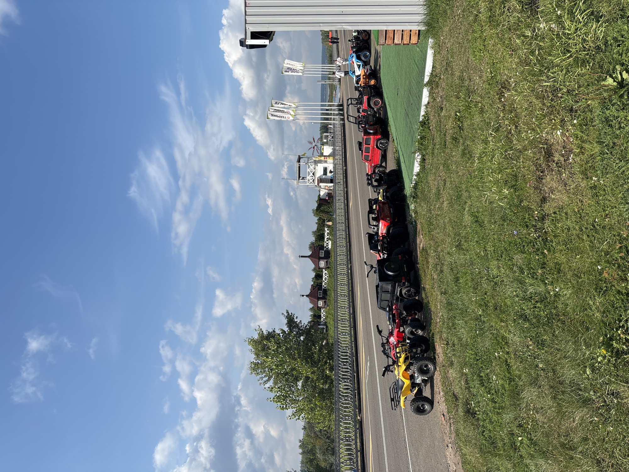 An exterior, eye-level, full shot captures a sunny day in Naberezhnye Chelny, Russia, with a clear blue sky dotted with white clouds. In the foreground, a line of ATVs and small off-road vehicles are parked on a green surface adjacent to a paved road. The vehicles are of various colors including yellow, red, black, orange, and blue. Behind the vehicles, a metal railing runs along the edge of a road, and beyond that, a large green tree stands prominently. In the middle ground, several red-roofed buildings with white trim are visible, resembling houses. Further in the distance, a white tower with a white structure on top, possibly a control tower or observation deck, is seen. Several white flags with text are planted in a line near the tower. A few people are visible walking in the background. The overall scene suggests an outdoor recreational area or a rental service for ATVs and similar vehicles.