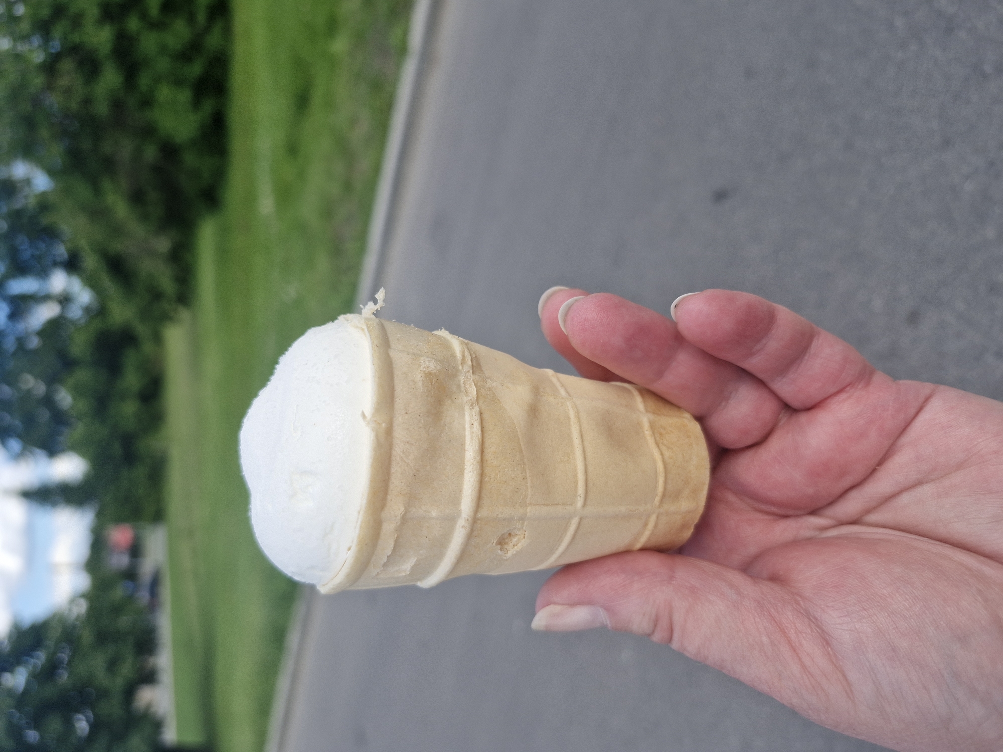 The image shows a close-up of a hand holding a partially eaten ice cream cone. The ice cream is white and appears to be vanilla flavored. The cone is a light golden brown color and has a waffle texture. The hand holding the cone has light skin and neatly trimmed fingernails. The background is blurred, but appears to be an outdoor setting with green grass and trees on the left, and a gray paved surface on the right, possibly a sidewalk or road. The lighting suggests it is daytime, and the overall scene is casual and relaxed, implying someone is enjoying a treat outdoors. There is no visible text in the image.