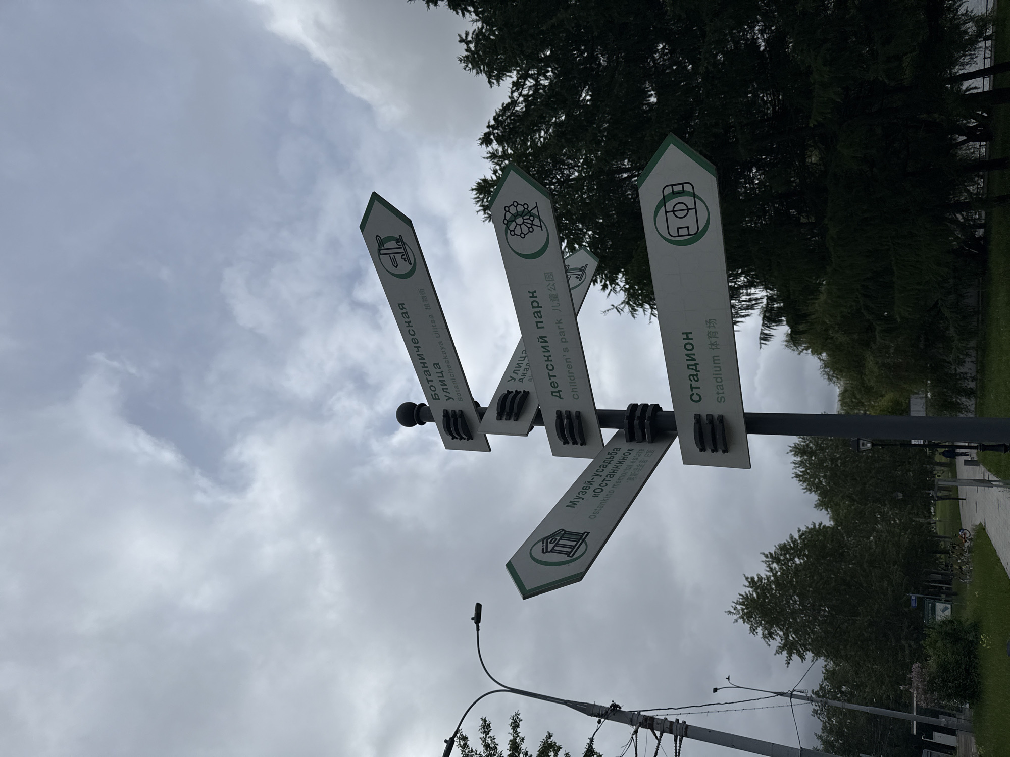The image shows a signpost with directional signs in multiple languages (Russian, English, and Chinese) in Unknown, Unknown. The signs point towards several locations: "Botanicheskaya Ulitsa (Botanical Street)," "Detskiy park (Children's park)," "Stadion (Stadium)," and "Музей-усадьба «Останкино» (Ostankino memorial estate)." The sky is cloudy, and there are trees in the background. The signpost is gray and has a black top. There are utility poles and wires in the lower part of the image.
