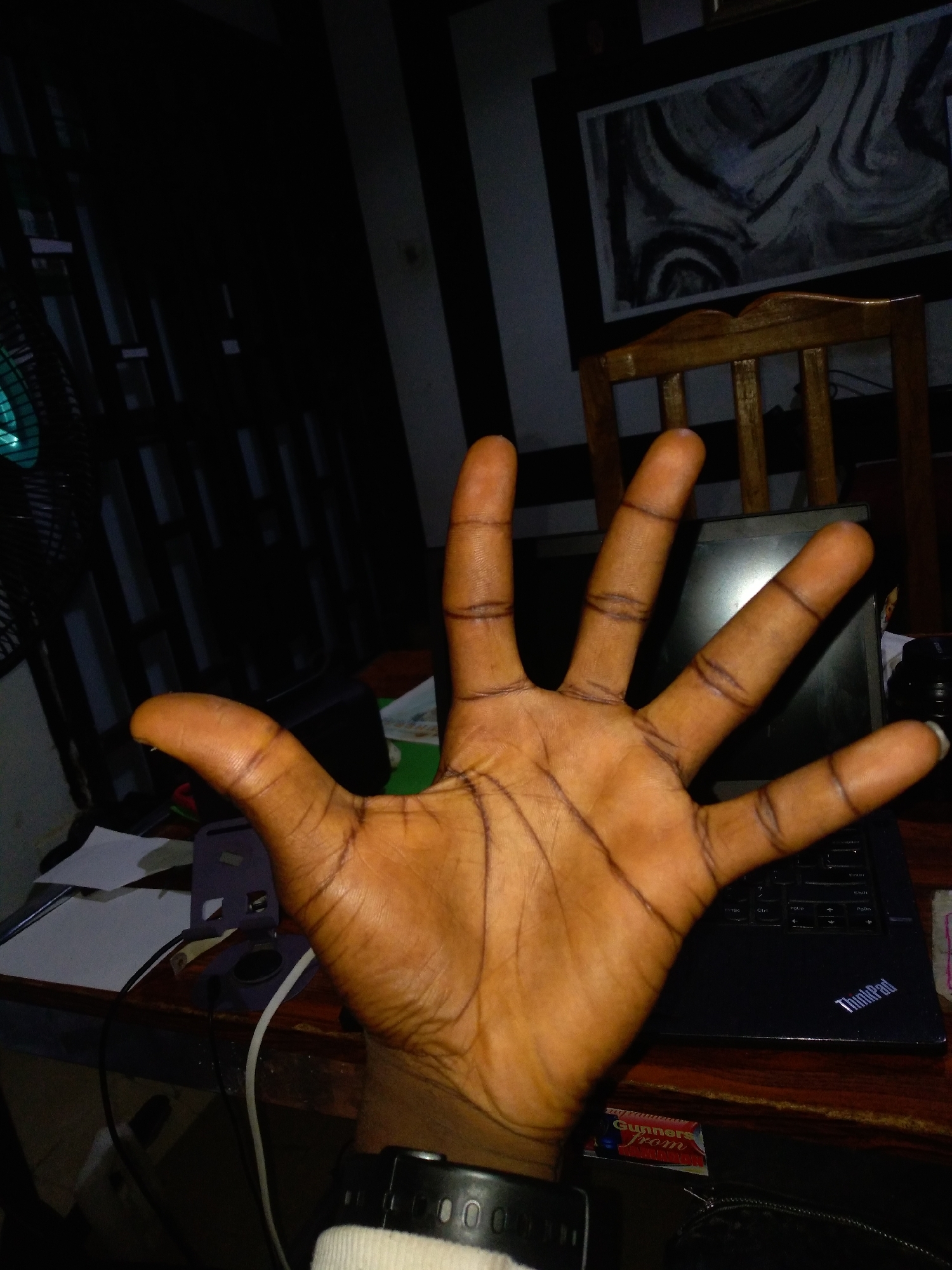 An over-the-shoulder shot captures a person's open hand displaying the palm with fingers extended upwards, against a dimly lit background. The hand is positioned in the foreground, with the palm facing the viewer and the fingers spread. The skin tone is a rich brown with visible lines and creases. The background is cluttered with objects suggesting a workspace or study area, including a laptop, papers, and a fan. The lighting is low, with harsh shadows cast by the fan. A wooden chair is visible behind the hand, and a framed artwork hangs on the wall. The location is indeterminate due to the lack of specific identifying features.