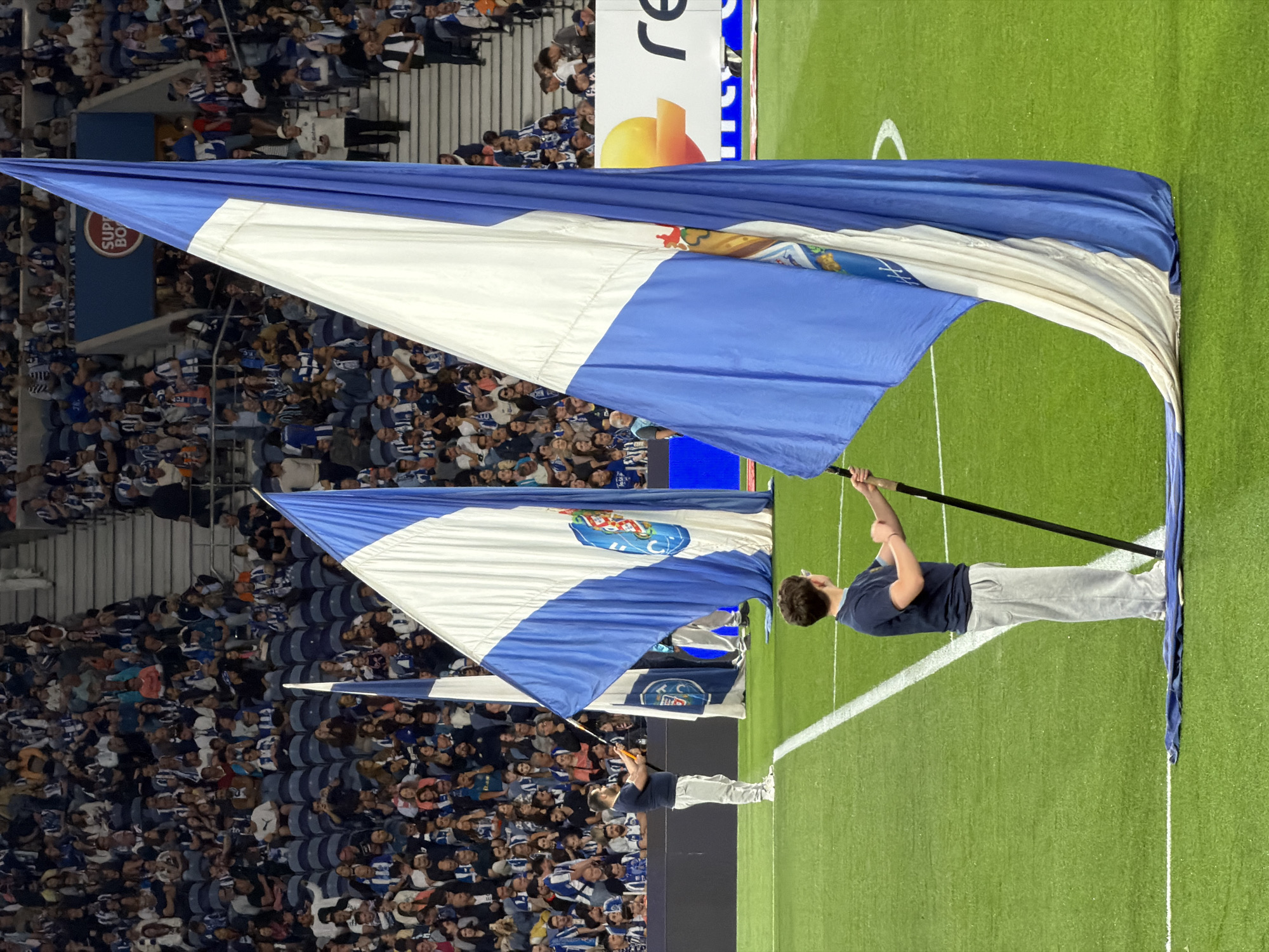 The image depicts a soccer match atmosphere, likely in Porto, Portugal, based on the presence of the FC Porto flag. A large, blue and white FC Porto flag is being held aloft on the edge of the field, close to the stands filled with fans. The fans are densely packed and mostly dressed in blue and white. Two individuals are holding flag poles. The field is green, with white lines marking the boundaries. A billboard featuring a partially obscured advertisement is visible behind the flag.