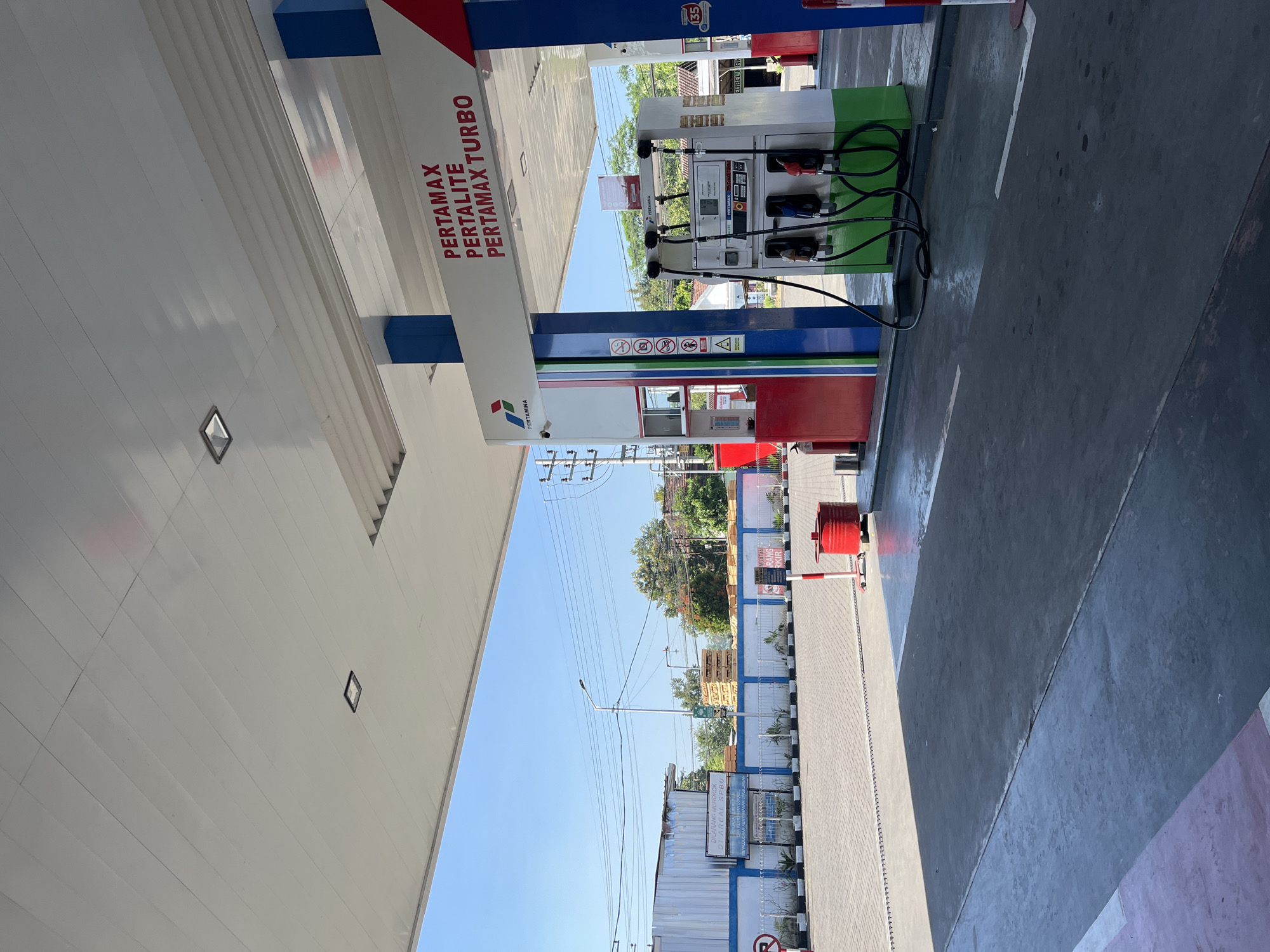 The image depicts a Pertamina gas station in Purworejo, Indonesia. The scene is captured from a low angle, looking up towards the canopy of the gas station. Under the canopy, a Pertamina gas pump is visible, with various fuel options like Pertamax, Pertalite, and Pertamax Turbo advertised. The pump has several nozzles ready for use. To the left of the pump, there is a red fire extinguisher. In the background, there is a paved area with a clear blue sky overhead. A white building with a blue stripe along the top is visible behind a low blue and white wall. There are also trees and power lines in the distance, suggesting an urban or suburban environment. The overall impression is one of a clean and functional service station.