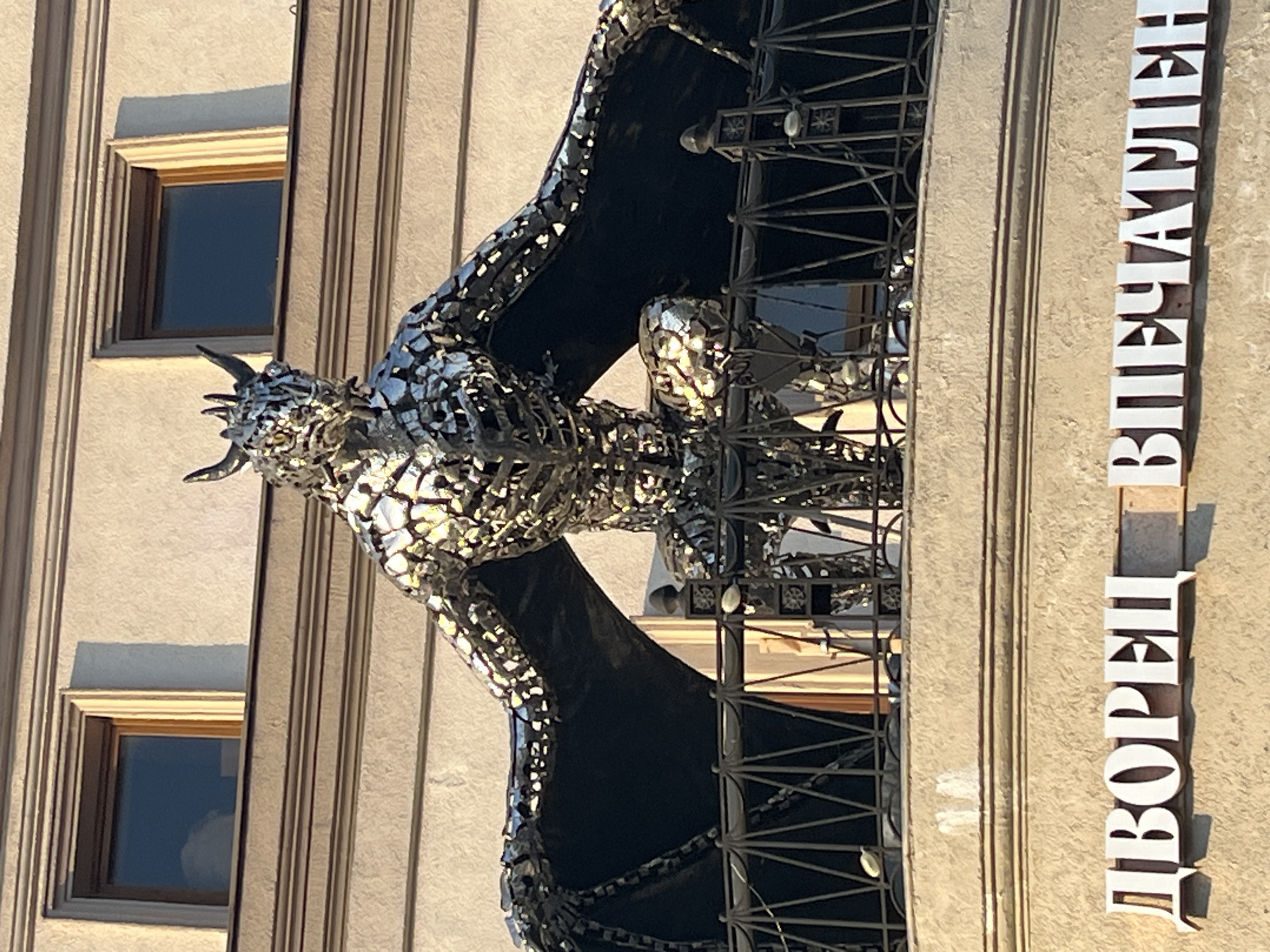 The image captures a metal sculpture of a dragon mounted on the side of a building. The building facade appears to be beige in color with recessed windows. The sculpture is made of shiny metal fragments with dark-colored wings. Next to the sculpture, a vertical sign in Cyrillic reads "ДВОРЕЦ ВПЕЧАТЛЕНИЙ," which translates to "Palace of Impressions" in English. This sign indicates that the building is likely the "Palace of Impressions" in Kazan, Russia. The photograph seems to be taken during daylight hours, with shadows cast on the building and sculpture.