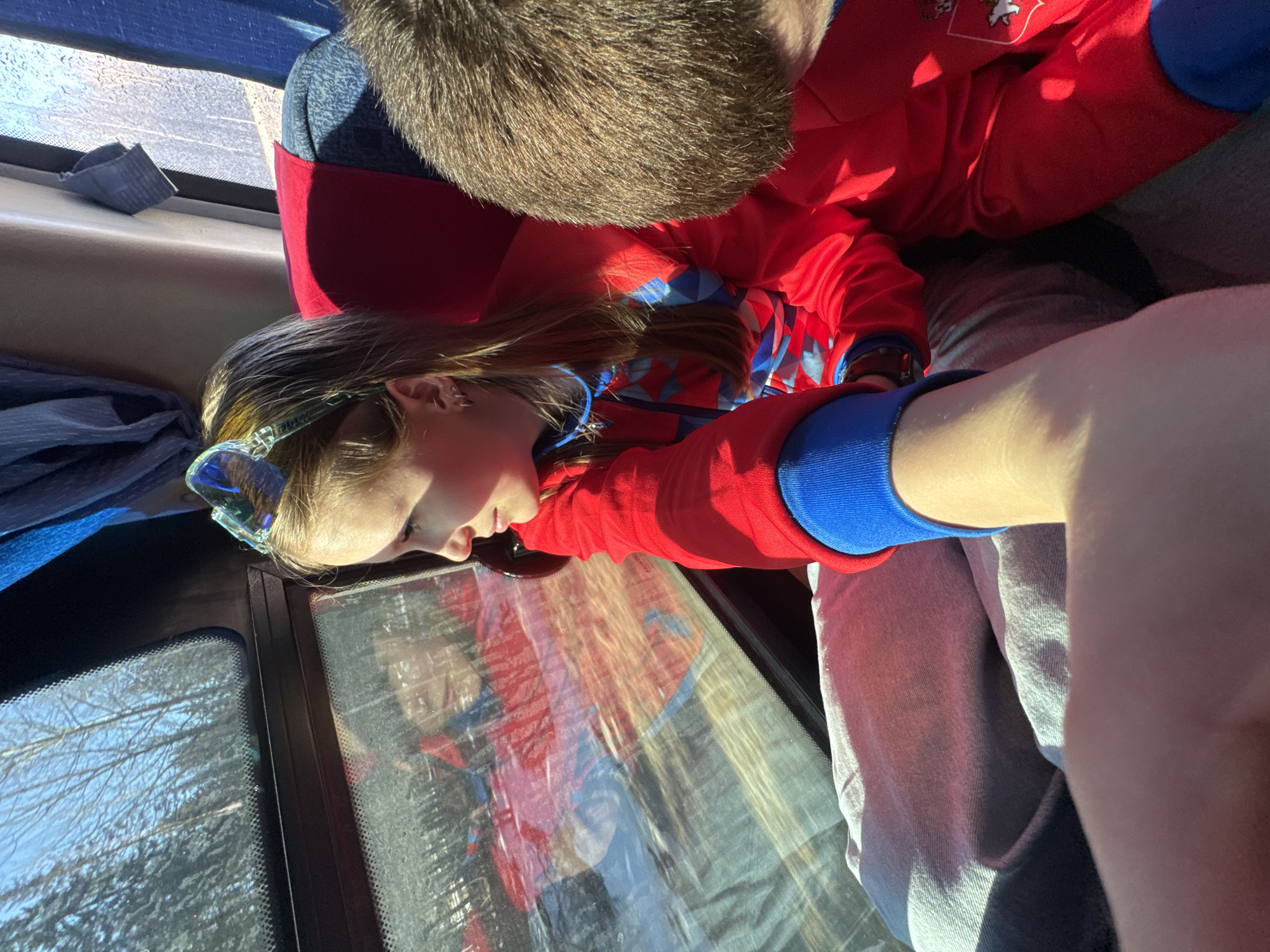 A young girl with light brown hair and a blue headband is looking out the window of a vehicle. She is wearing a red and blue outfit. A person with short brown hair is beside her, also wearing a red and blue outfit. The window shows reflections of the people in the car and trees. The exact location in the city of Челва, Russia, cannot be determined from the image.