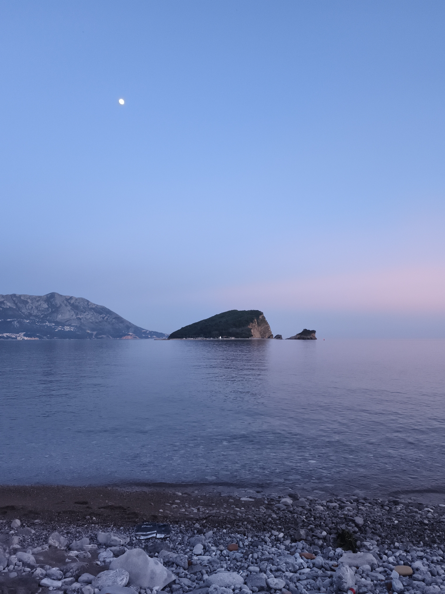 The image displays a tranquil coastal landscape captured during twilight, either at dawn or dusk. The sky is a clear, gradient blend, transitioning from a light blue overhead to soft hues of pale pink and purple near the horizon on the right. A distinct, white moon, appearing as a crescent or gibbous shape, is visible in the upper left portion of the sky.
A calm body of water, likely the sea, fills the mid-ground, subtly reflecting the sky's colors. In the distant background to the left, rugged, grey mountains rise, with scattered lights indicating human habitation along their lower slopes. Prominently featured in the mid-ground is a verdant island or large rock formation densely covered in green foliage. To its right, a smaller, bare rock formation protrudes from the water, accompanied by a small red buoy.
The foreground is a wide, pebbled beach composed of various sizes of smooth, grey, and white stones. Among these pebbles, in the lower left quadrant, a black object resembling a plastic bag is visible. The text "ASTORIA" and partially "VIBER..." can be clearly read on this object.
The scene conveys a peaceful and serene atmosphere, with no people or overt activity present. The clear sky and calm water suggest pleasant weather conditions. This picturesque setting is consistent with coastal regions, specifically Budva, Montenegro, as indicated by the location context.