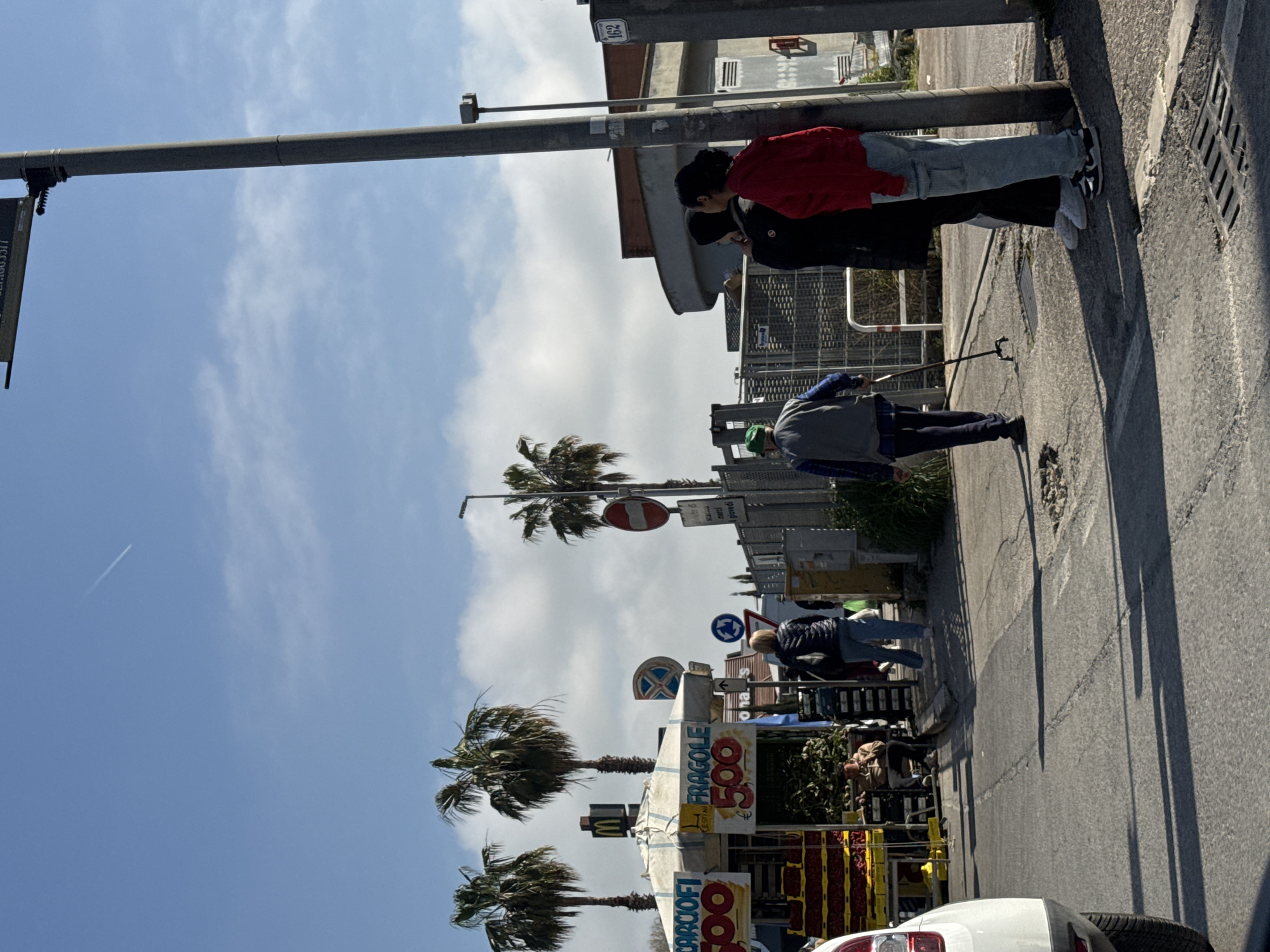 The image shows a street scene in Nettuno, Italy. Two individuals are walking on the sidewalk near the edge of the road. One person uses a walking stick. A street vendor displaying fruit appears to be in business on the side of the road. There are palm trees along the side of the road. There are several street signs. The sky is blue and partly cloudy.
