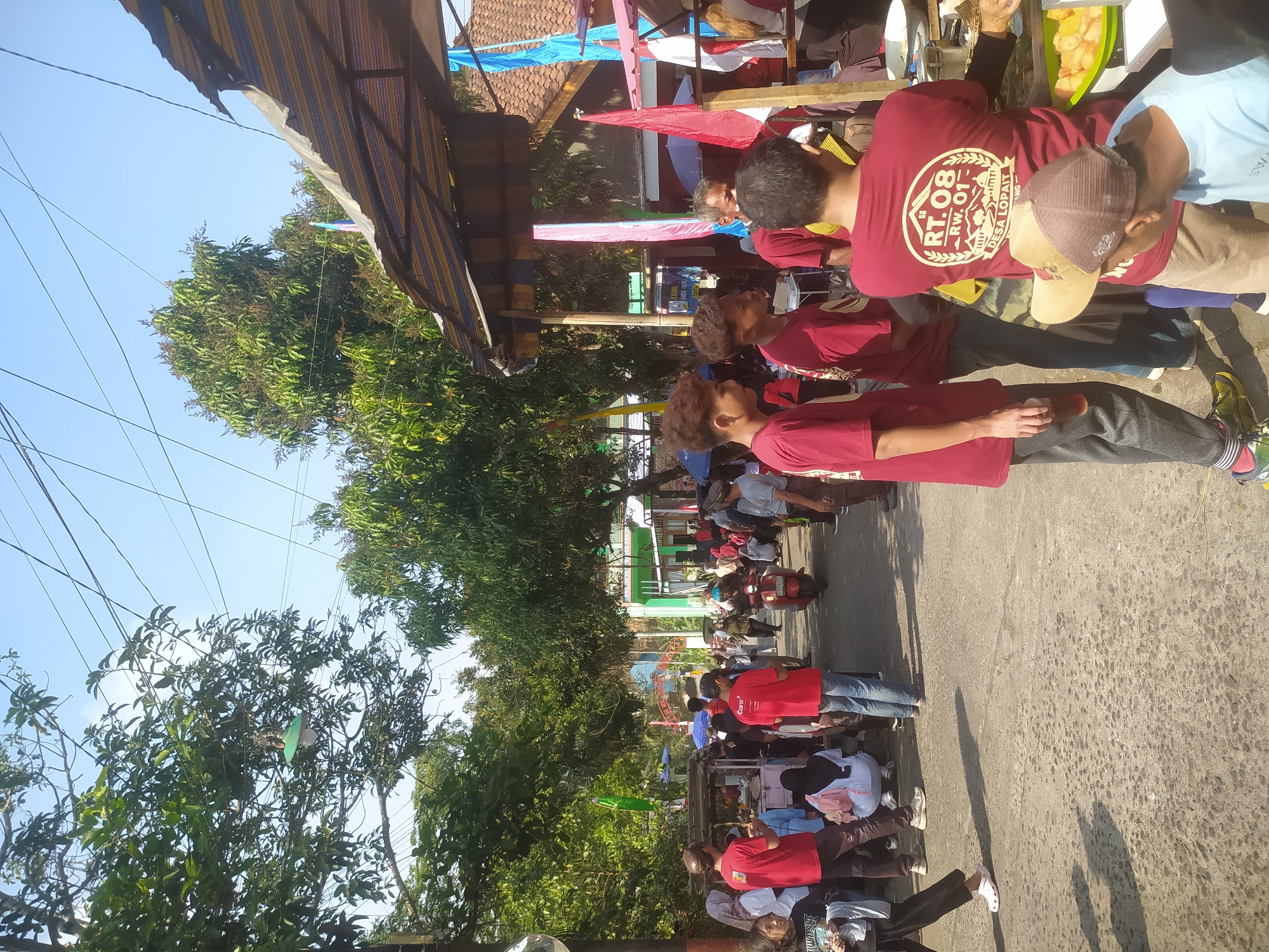 The image depicts an outdoor scene in the city of Lopait, Indonesia, likely during a daytime event or gathering. A street is lined with trees on the left and stalls or temporary structures on the right. A group of people, predominantly wearing red t-shirts, are congregated on the street and near the stalls.
In the foreground, several individuals are visible. On the left, a person in a black shirt and black pants with white Crocs walks away from the camera. To their right, a person in a red t-shirt is holding a plastic cup with a dark liquid. Behind them, several other individuals in red t-shirts stand and crouch. Two young males in red t-shirts are looking to their left, and another person in a red t-shirt with a graphic design on the back is facing away from the camera. A man with a brown cap is also in the foreground, looking down.
In the mid-ground, a scooter with a rider passes through the street. More people are visible milling around, some under colorful awnings and umbrellas. Flags and banners, some appearing to be red and white, are displayed. Stalls are set up, with food or goods displayed on tables.
The environment includes a paved street, trees with lush green foliage, and structures with corrugated roofs. The sky is clear and blue. Power lines are visible overhead. The overall impression is a lively community event. The t-shirt on one of the individuals clearly displays "RT.08 RW.01 DESA LOPAIT", confirming the location.