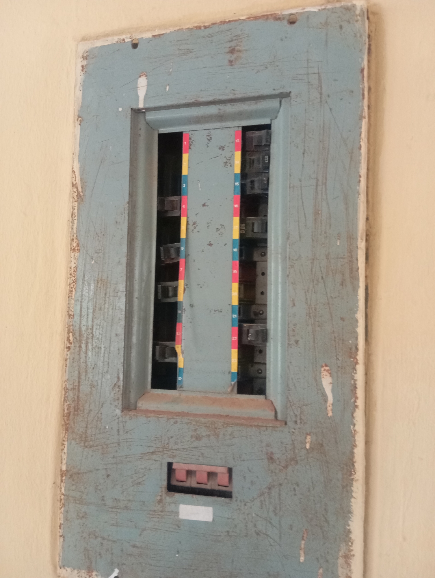The image shows a close-up, head-on view of an old, weathered, blue metal electrical panel mounted on a cream-colored wall. The panel has visible scratches and rust marks, indicating age and exposure. A hinged door, slightly ajar, reveals a row of circuit breakers within. Colored vertical strips, likely indicating breaker numbers or types, are visible to the left of the breakers, with numbers like "13", "15", "16", "18", "19", "21", and "22" partially visible. Below the breaker compartment, there is a small, rectangular panel with three pink toggle switches. A white label is affixed below this switch panel. The lighting is neutral, suggesting an indoor setting during daylight hours. The image is taken in Jos, Nigeria.