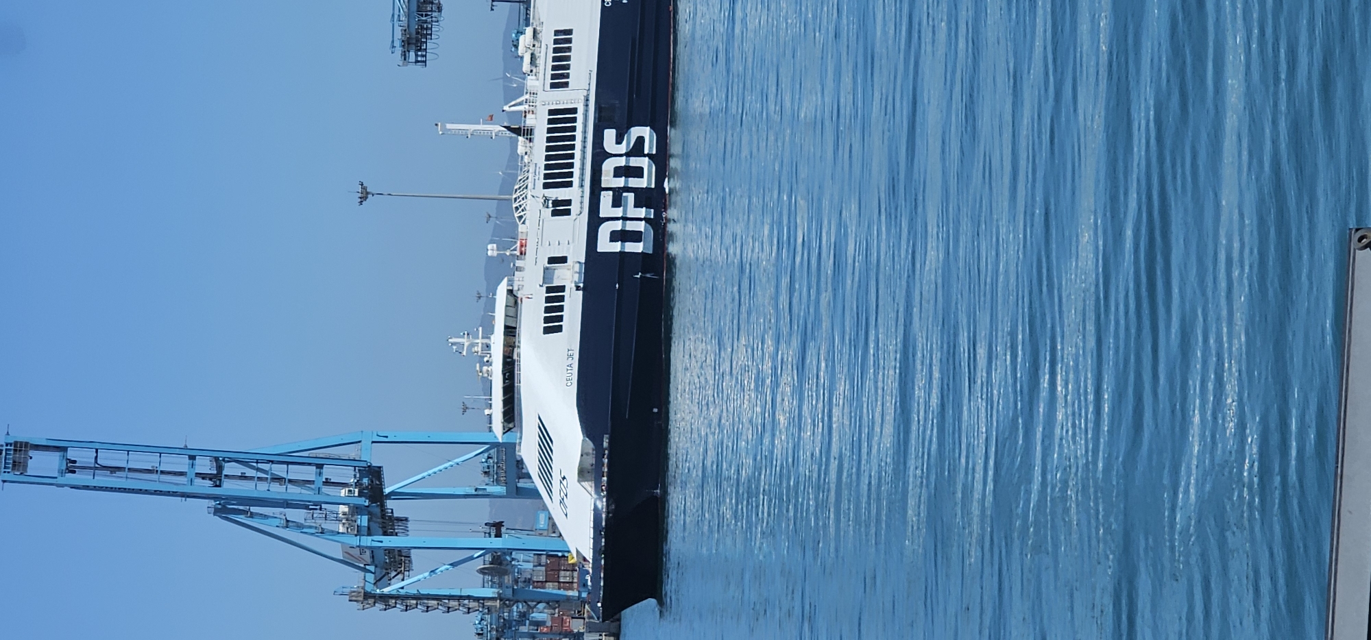 This media file captures a large vessel, identified as a ferry or roll-on/roll-off ship, in a port setting. The ship is primarily black and white, featuring the prominent logo "DFDS" in white letters on its side. Below the logo and further towards the bow, the text "CELTA JET" is also visible.
The vessel is situated on calm, blue water, which shows subtle ripples. In the background, to the left of the ship, several large, bright blue port cranes are visible, indicative of a busy shipping terminal. Stacks of shipping containers can be discerned beneath and around the cranes. The sky above is clear and a vibrant blue, suggesting bright daylight and good weather.
The scene depicts an active maritime environment, likely a commercial port. Given the context, this port is Algeciras, Spain, a major hub for shipping and ferry services. The image focuses on the ship and the industrial infrastructure of the port under clear skies.