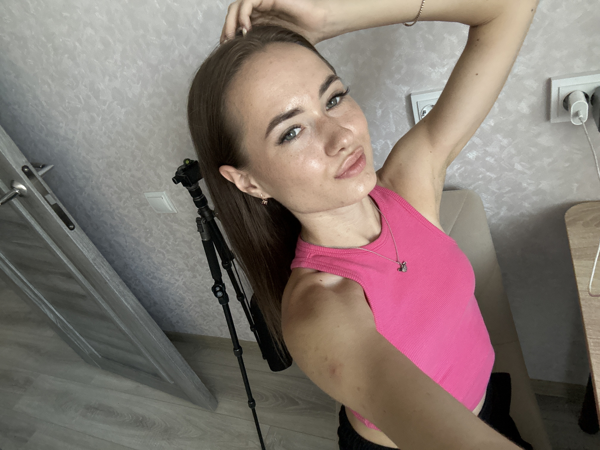 In a domestic indoor setting, a young woman with long brown hair and light green eyes poses for a selfie. She is wearing a pink ribbed tank top and has her left hand touching her hair. The background features a textured light gray wall, a door frame to the left, a tripod with a camera attached, and electrical outlets on the wall. To the right, part of a beige chair and a wooden surface are visible. The lighting is bright, likely from an artificial source.