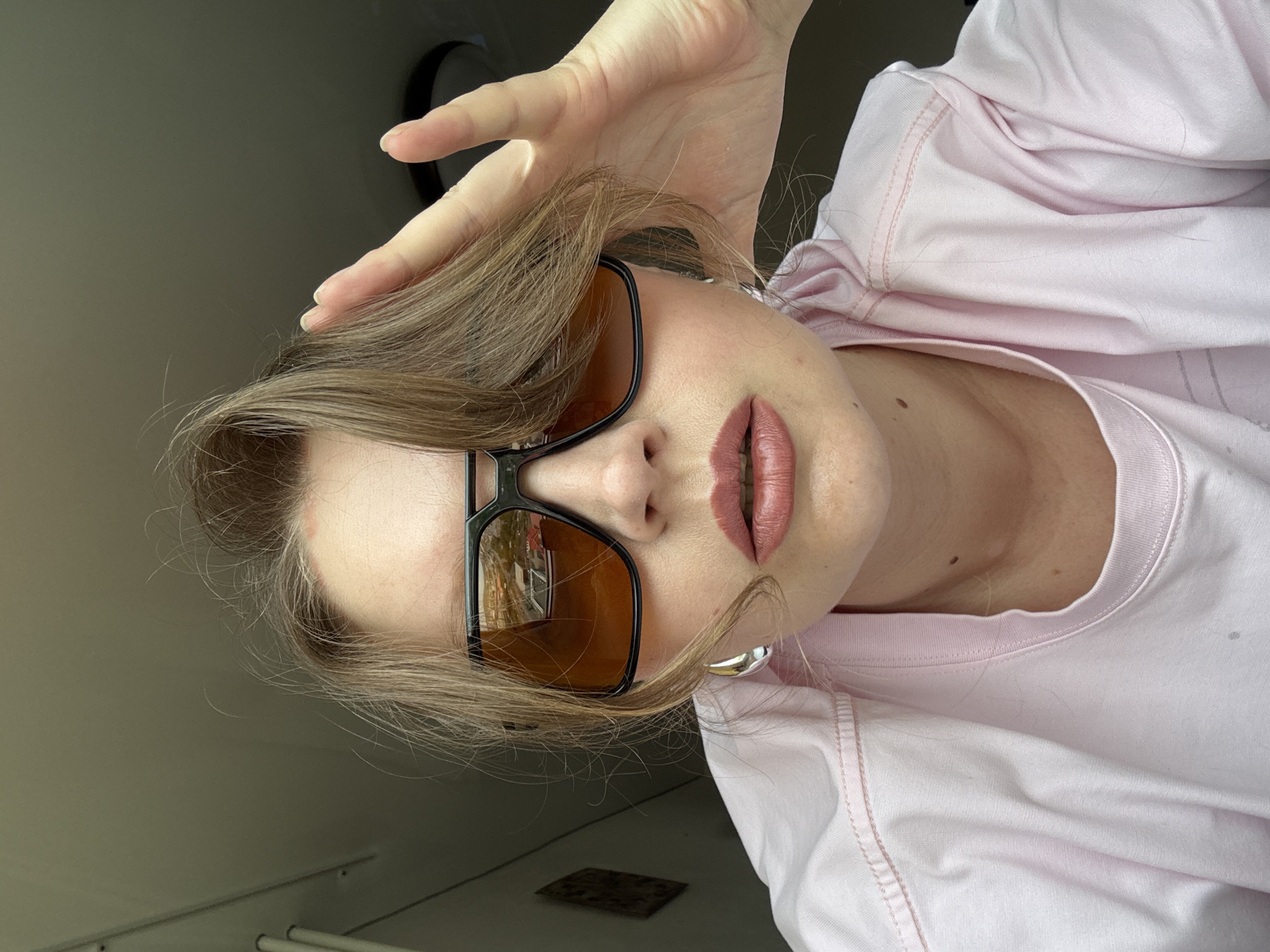A woman poses, wearing a light pink t-shirt and large black-framed sunglasses with orange lenses. Her hair is light brown and she is holding it with her right hand. She is wearing silver hoop earrings. The reflection in the sunglasses suggests an urban environment, although the specific location within Izhevsk, Russia cannot be determined from the image alone.