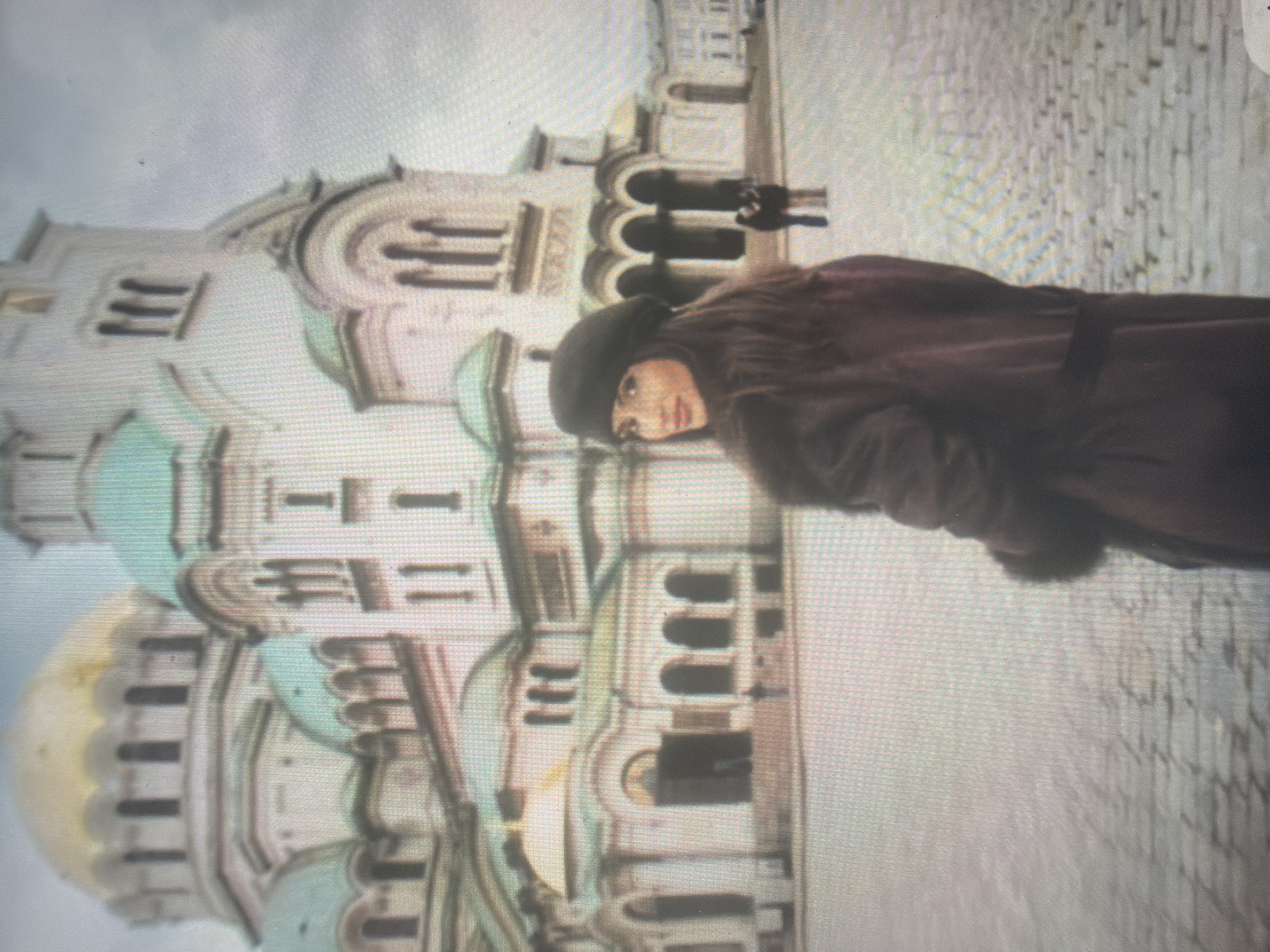 The image depicts an outdoor scene in front of a grand Eastern Orthodox cathedral in Shumen, Bulgaria, under an overcast sky. In the foreground, positioned slightly right of center, a woman with long, dark, wavy hair and a dark beret is captured looking back over her left shoulder towards the camera. She has visible makeup, including lipstick, and exhibits a slight smile. She is wearing a dark, possibly fur-trimmed, long coat in a color resembling dark brown or maroon. The ground beneath her and extending into the mid-ground is paved with light-colored cobblestones.
The dominant background element is a large, multi-tiered cathedral featuring a prominent central golden dome and numerous smaller teal-colored domes and roofs. The structure is constructed from light-colored stone or stucco and exhibits intricate architectural details including arched windows, multiple entrances with arches, and decorative elements. In the mid-distance, to the right of the main cathedral entrance, two additional figures are visible standing near the building. The sky is uniformly gray, indicating an overcast day with soft, diffused lighting.