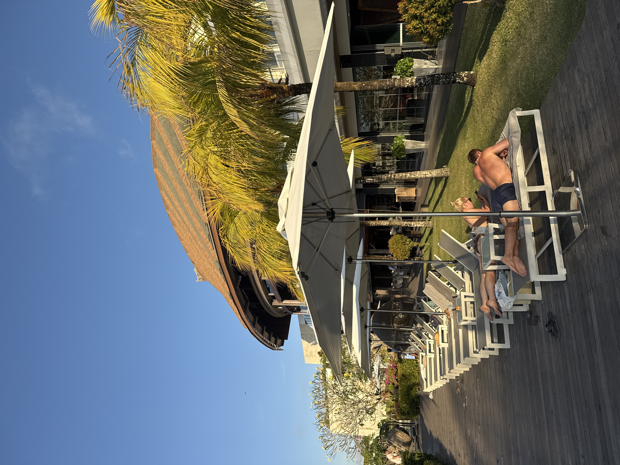 A group of people are relaxing on lounge chairs in front of a building with a curved, thatched roof. The scene appears to be a resort in Kuta Selatan, Indonesia, given the tropical vegetation, palm trees, and clear blue sky. Two individuals are prominently featured in the foreground, a man and a woman, both lying on lounge chairs. The man is on his stomach, facing away from the camera, and the woman is on her side, looking at her phone. In the background, several other lounge chairs are visible, some occupied and some empty, lined up along a wooden deck. Large white umbrellas are strategically placed to provide shade. The building itself is modern, with large glass windows and a grey facade, contrasting with the natural elements surrounding it. The overall atmosphere is one of leisure and relaxation, bathed in warm sunlight.