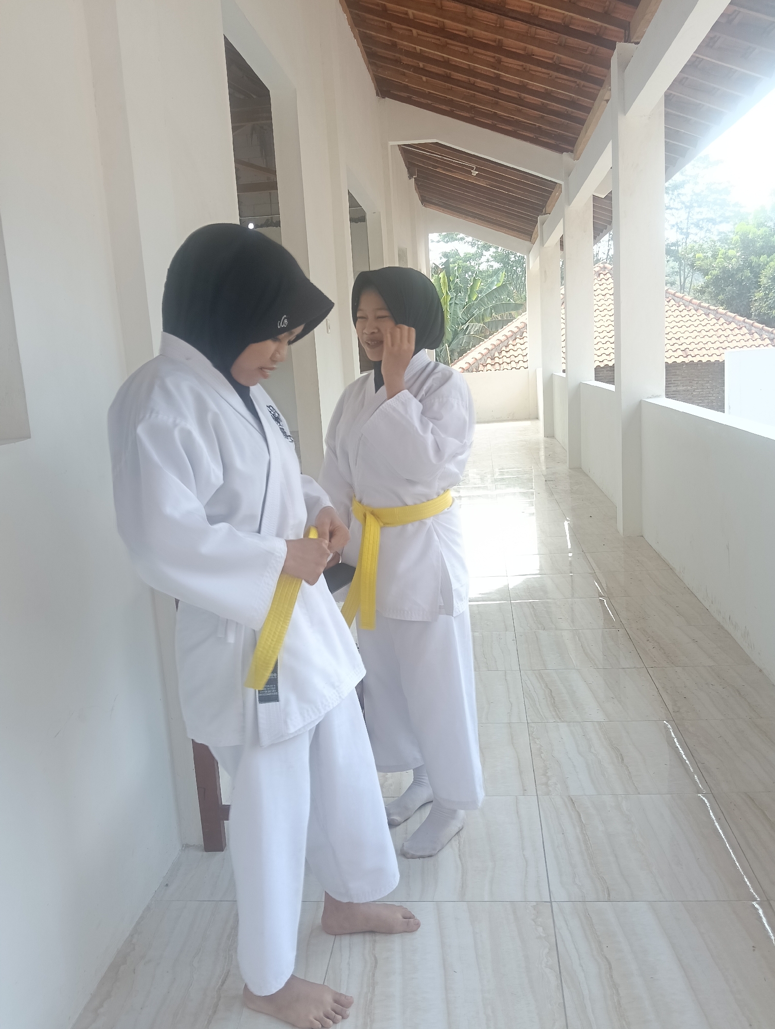 Two young women in white karate uniforms with yellow belts stand on a tiled outdoor walkway. The woman on the left, wearing a black hijab, is adjusting her yellow belt. The woman on the right, also wearing a black hijab, is smiling and touching her face. The walkway is covered by a pitched roof supported by white pillars. Beyond the walkway, tiled roofs and greenery are visible. The scene appears to be outdoors in Adinuso, Indonesia, likely a school or community center with martial arts facilities.
