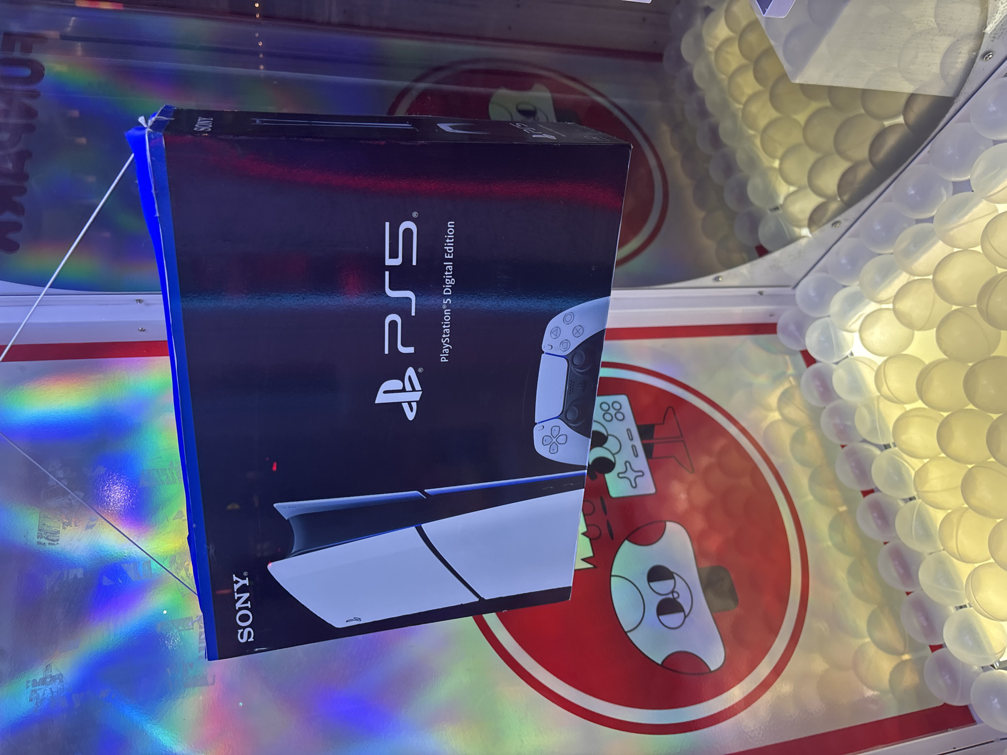 A PlayStation 5 Digital Edition console box is prominently displayed in a vertical orientation within what appears to be an arcade game or claw machine. The box, predominantly black, features "PS5" in large white lettering, with "PlayStation®5 Digital Edition" and the Sony logo clearly visible. An image of the console itself and a DualSense controller are also printed on the box.
The box is held in place by a blue acrylic or plastic bracket at the top, from which a thin white string or wire extends. Below and to the left of the box, a surface exhibits vibrant holographic, iridescent reflections, creating rainbow patterns. On this surface, a red circular logo with a cartoon-like character holding a game controller is visible. The character has an "88" and controller button symbols printed on it.
To the right of the PS5 box, a wall of translucent white or off-white plastic spheres or capsules, commonly found in prize machines, fills the frame. The artificial lighting inside the machine casts soft shadows among these spheres. The overall setting is an indoor entertainment venue, likely an arcade or game center, located in Buenos Aires, Argentina, designed to attract players to win the featured prize.