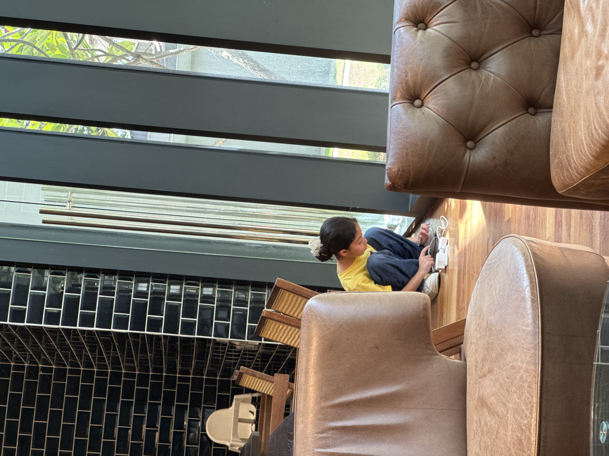 In a modern cafe with dark tiled walls and a large window featuring vertical grey panels, a young girl with her hair in a bun adorned with a white flower is kneeling on the wooden floor. She is wearing a yellow t-shirt and blue jeans, and she appears to be interacting with a white mobile device and its charger. Two plush brown leather chairs are prominently placed in the foreground, partially obscuring the view of the girl. In the background, high chairs and wooden chairs with woven seats are visible. Through the window, greenery and trees can be seen, suggesting an outdoor environment. The scene is lit by natural light streaming in from the window, casting shadows on the floor. The cafe appears to be located in Kuala Lumpur, Malaysia, based on the provided context.