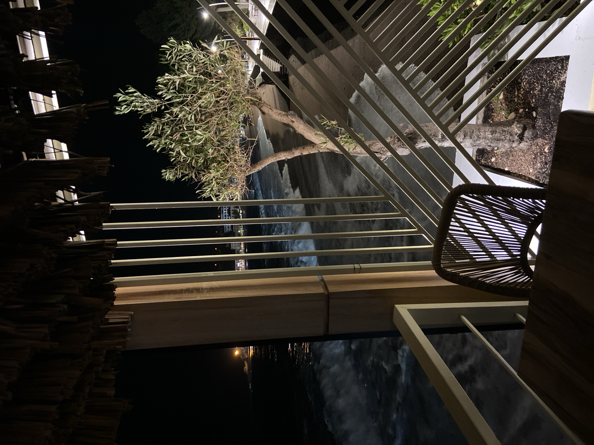 At night, the camera looks out from a balcony in Petrovac centar, Montenegro. Below is water. To the left, a thatched roof is visible. In the distance, lights illuminate the shoreline. In front of the water, a tree grows out of a planter. A metal railing partially obscures the view. A chair and table are on the balcony.