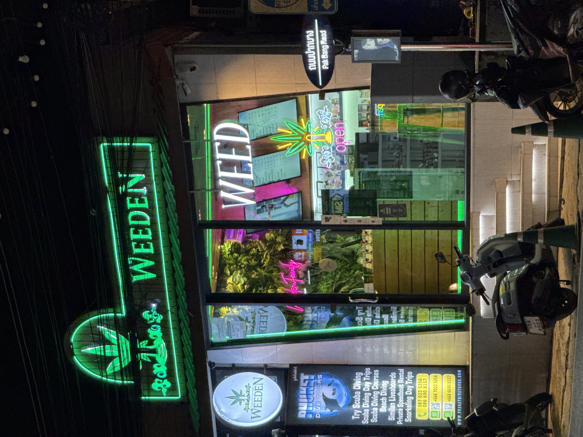 The image shows a brightly lit cannabis dispensary named "WEED" in Karon, Thailand, at night. The shop's facade features a large green neon sign with a cannabis leaf motif and the word "WEED" illuminated in green. A circular sign on the left side of the entrance also displays the "WEEDEN" logo. Inside, lush green plants are visible through the large glass windows, and shelves are stocked with products. A pink neon sign spelling "Weeden Family" is also visible inside.
In front of the shop, two scooters are parked. One is a light blue scooter with a Thai license plate that reads "1กผ 3923" and a white helmet on the seat. The other is a darker scooter partially visible on the left. A green traffic cone stands near the blue scooter.
To the right of the entrance, a dark blue sign with white text indicates "ถนนปากบาง Pak Bang Road". There's also a black and white image of a person on a poster attached to a pole. The street is paved, and the building is lit by streetlights and the shop's neon signs. The environment suggests a commercial street in a tropical location, possibly a tourist area given the "Phuket Dive Tours" advertisement on the left side of the entrance.